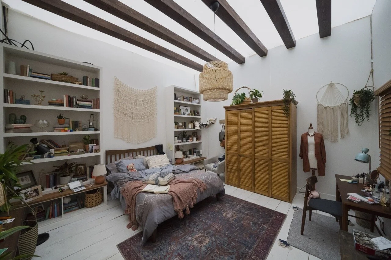 A cozy bedroom with white walls, dark wooden ceiling beams, and a white wooden floor. There is a bed with gray bedding and a pink blanket, next to two white bookshelves filled with books and decor. A large, woven wall hanging is behind the bed. A wooden wardrobe is on the right, with potted plants on top. A mannequin with a brown sweater is near a wall decorated with macrame wall hangings. A small desk with a blue lamp and various items is on the far right, and a wooden chair is placed at the desk. A patterned rug covers the floor in front of the bed.