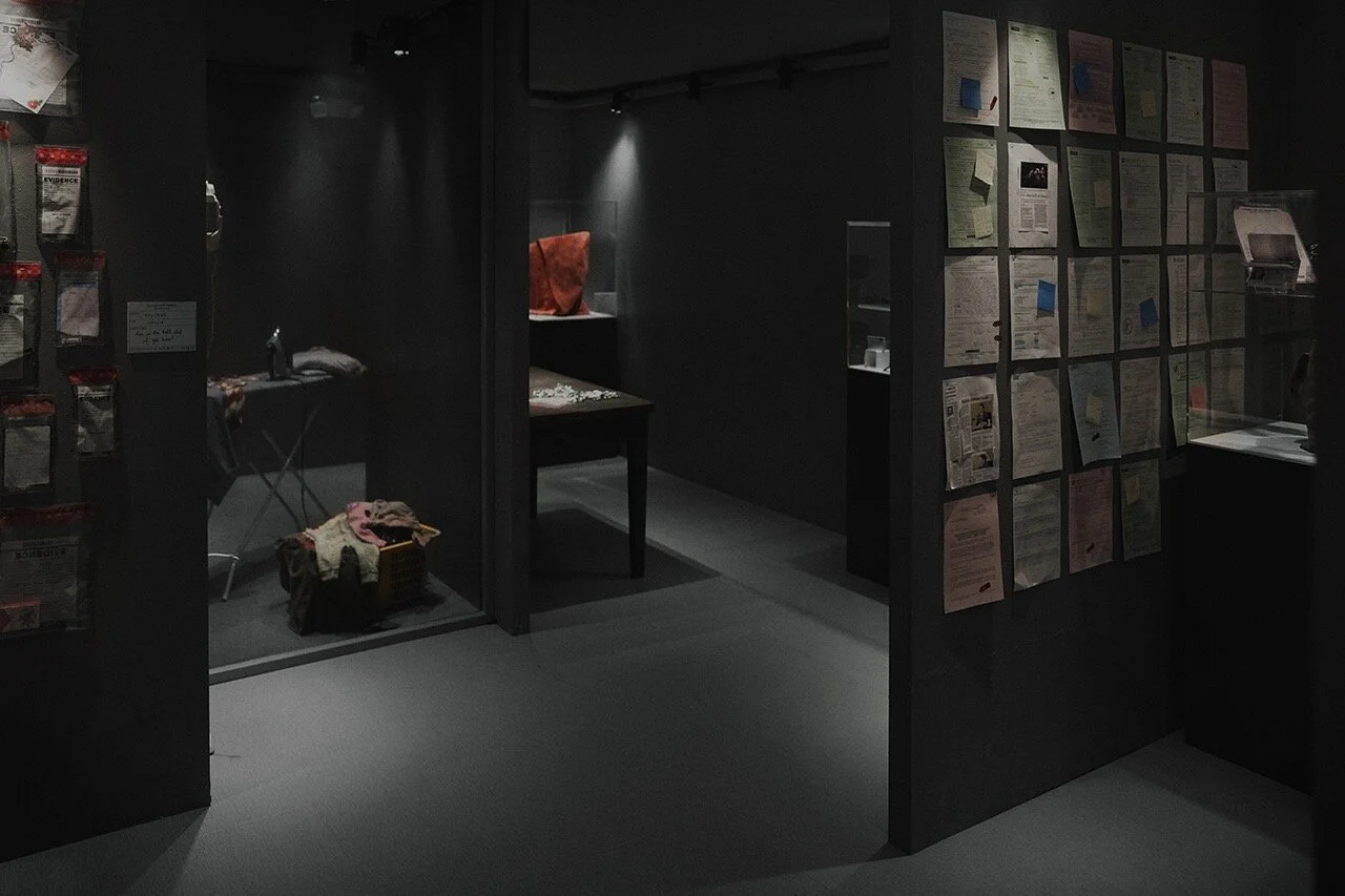 Dark exhibition room with display cases, charts on the wall, and clothing items on a rack.