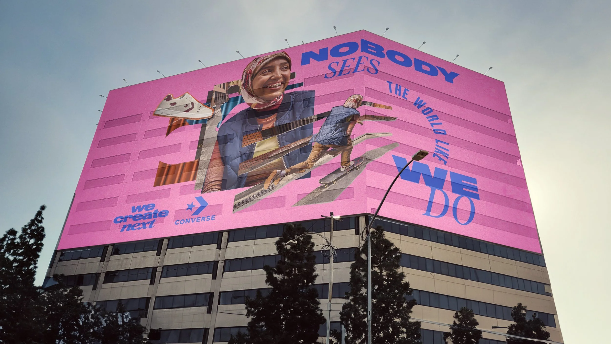 Large pink billboard on a building featuring a smiling woman with a headscarf, skateboarding, and wearing casual clothes. The billboard has the words 'Nobody Sells the World Like We Do' and mentions the brands 'Converse' and 'Next'.