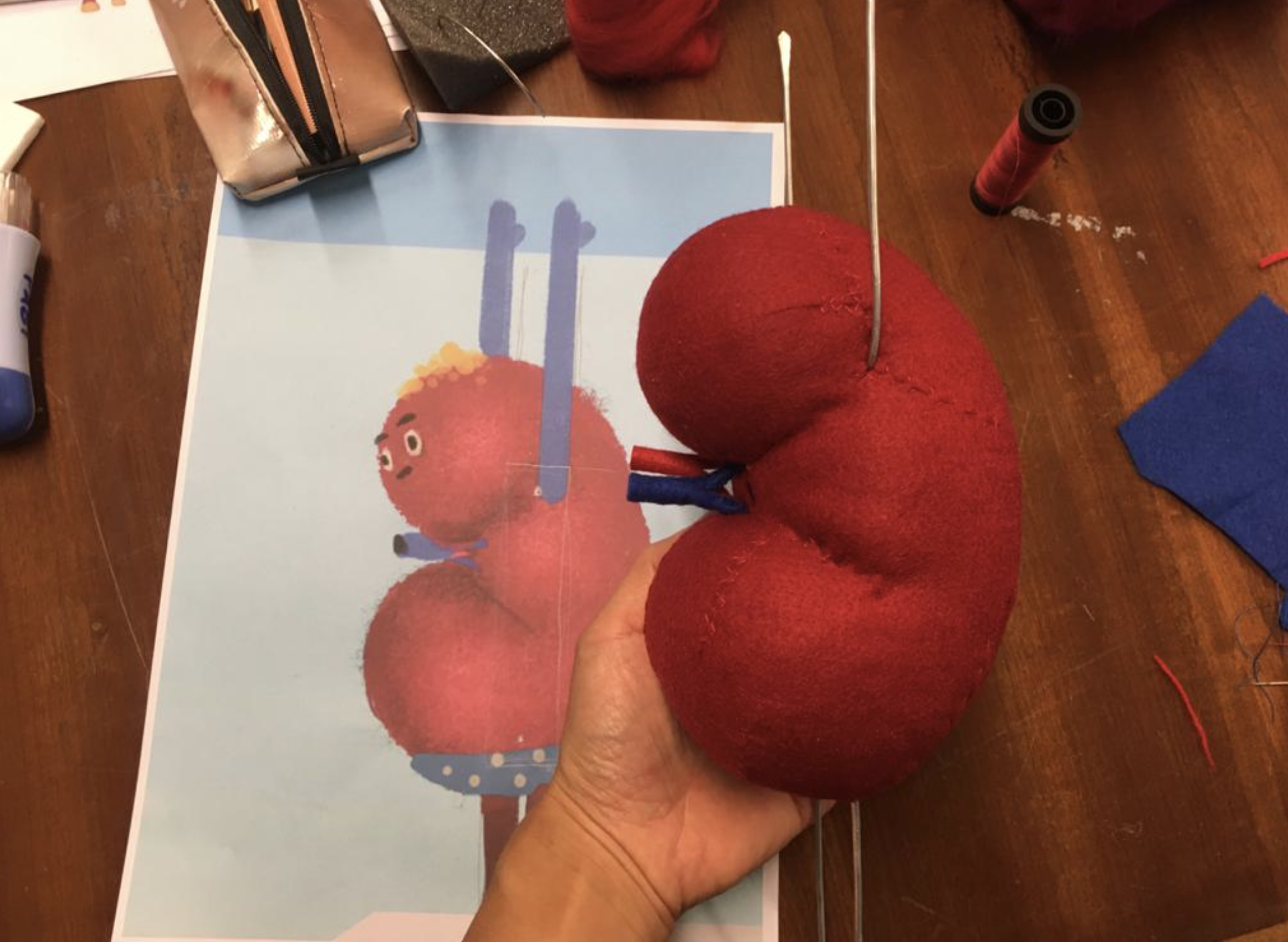 A craft project with two large red apples made of felt, with a hand holding them, placed on a wooden table. There are art supplies and a printed illustration of a character with curly hair and red apples on the table.