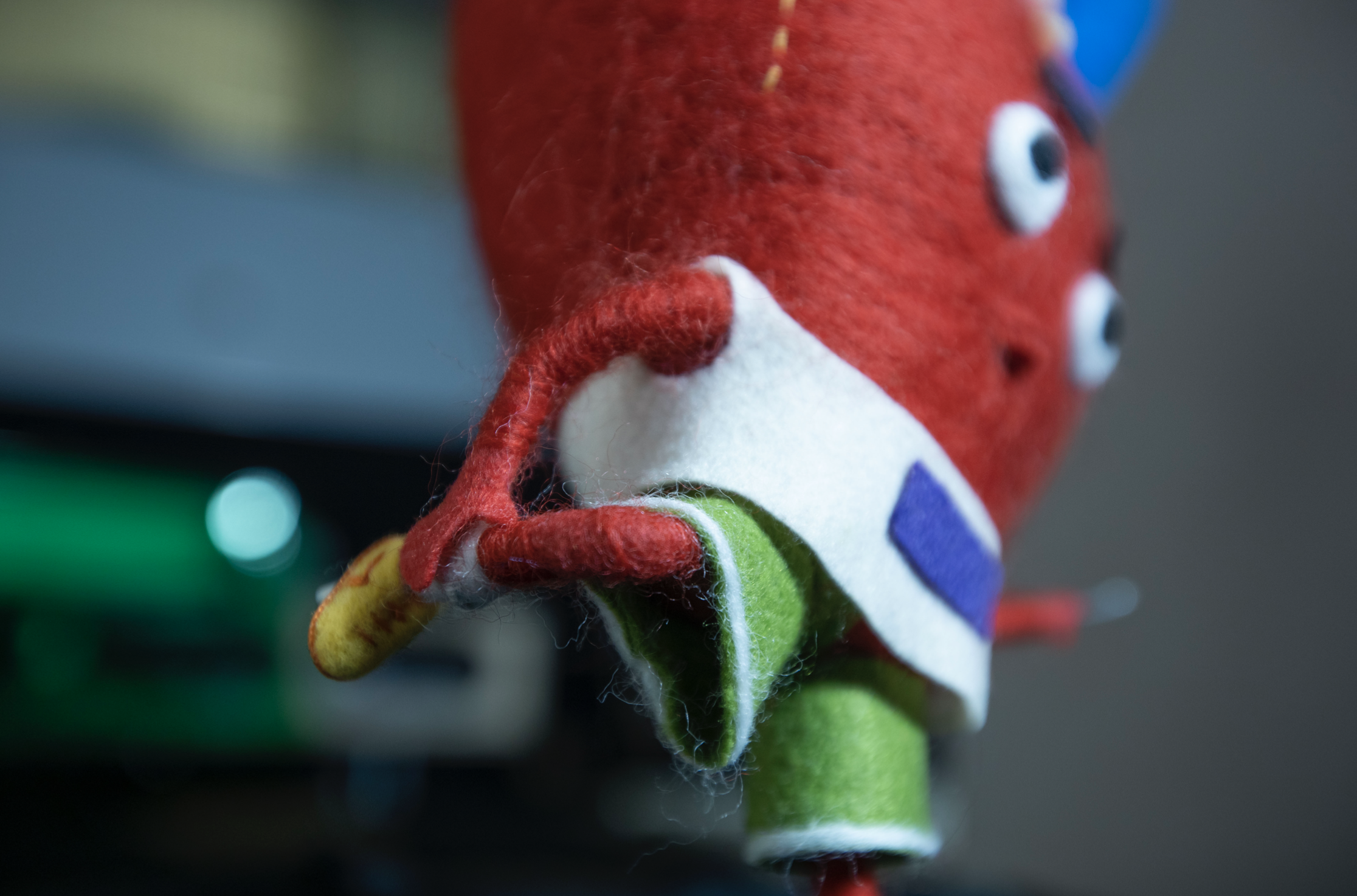 Close-up of a red felt plush toy with eyes, wearing a white, blue, and green costume, with a yellow accessory on its arm.