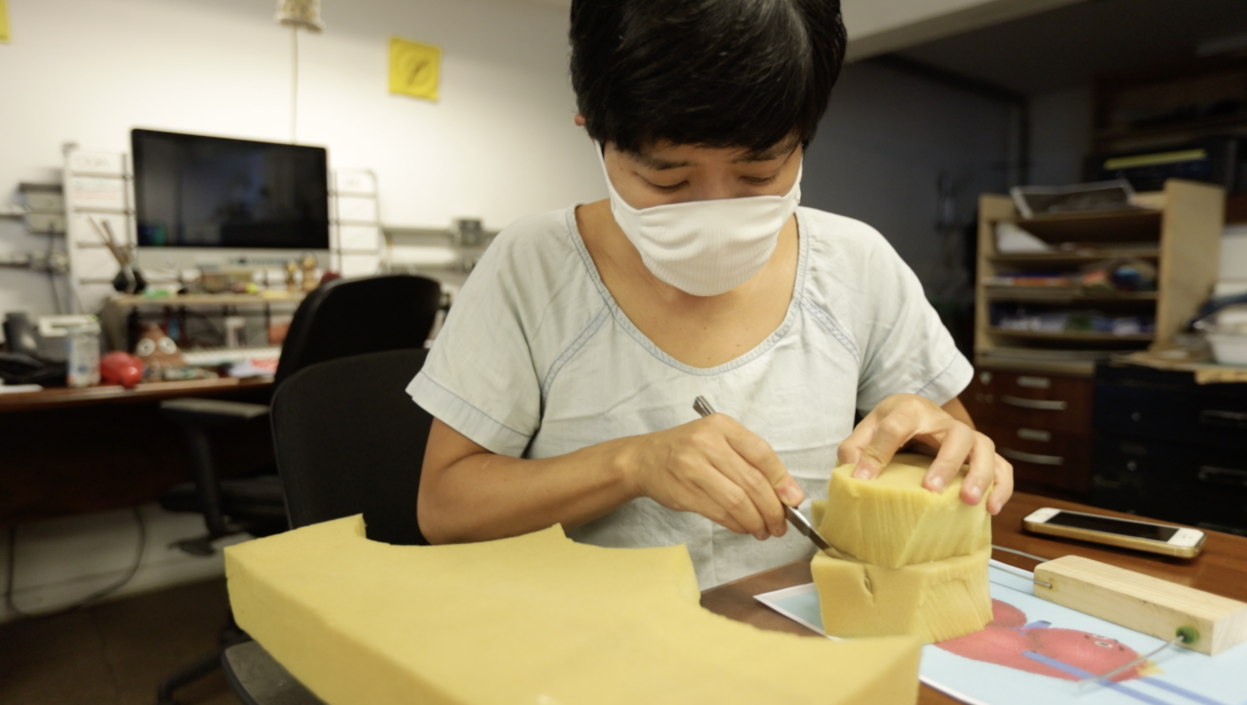 Person wearing a face mask and a light-colored shirt engaged in a craft or project involving yellow foam or sponge, sitting at a table cluttered with various items, in an indoor workspace or office.