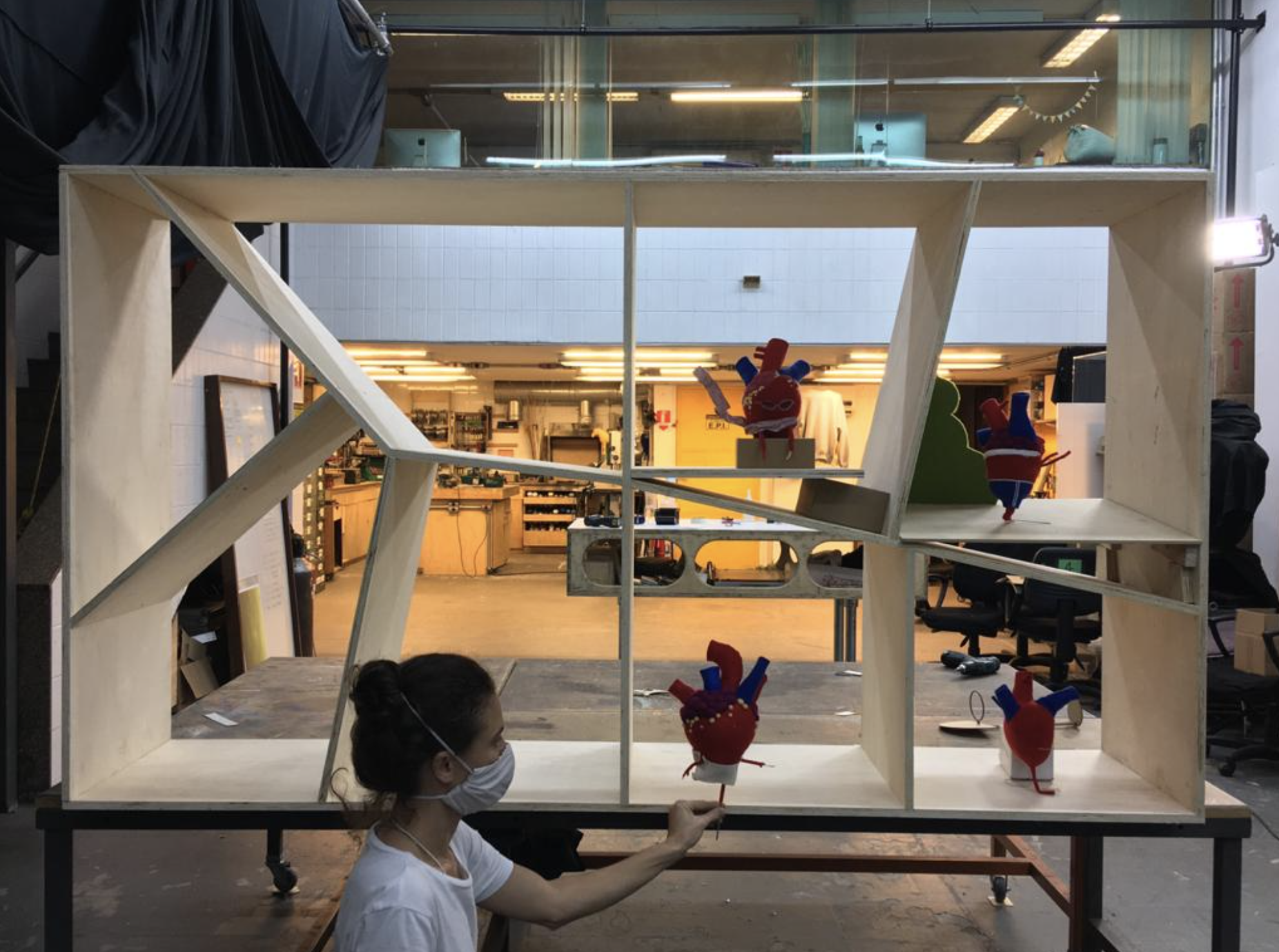 A woman with a face mask is holding a puppet of a heart with arms and legs, positioned in front of a large wooden structure with multiple compartments. Inside the compartments are several other similar heart puppets, some with limbs and clothing accessories, displayed as part of an art or design setup in a workshop or studio environment.