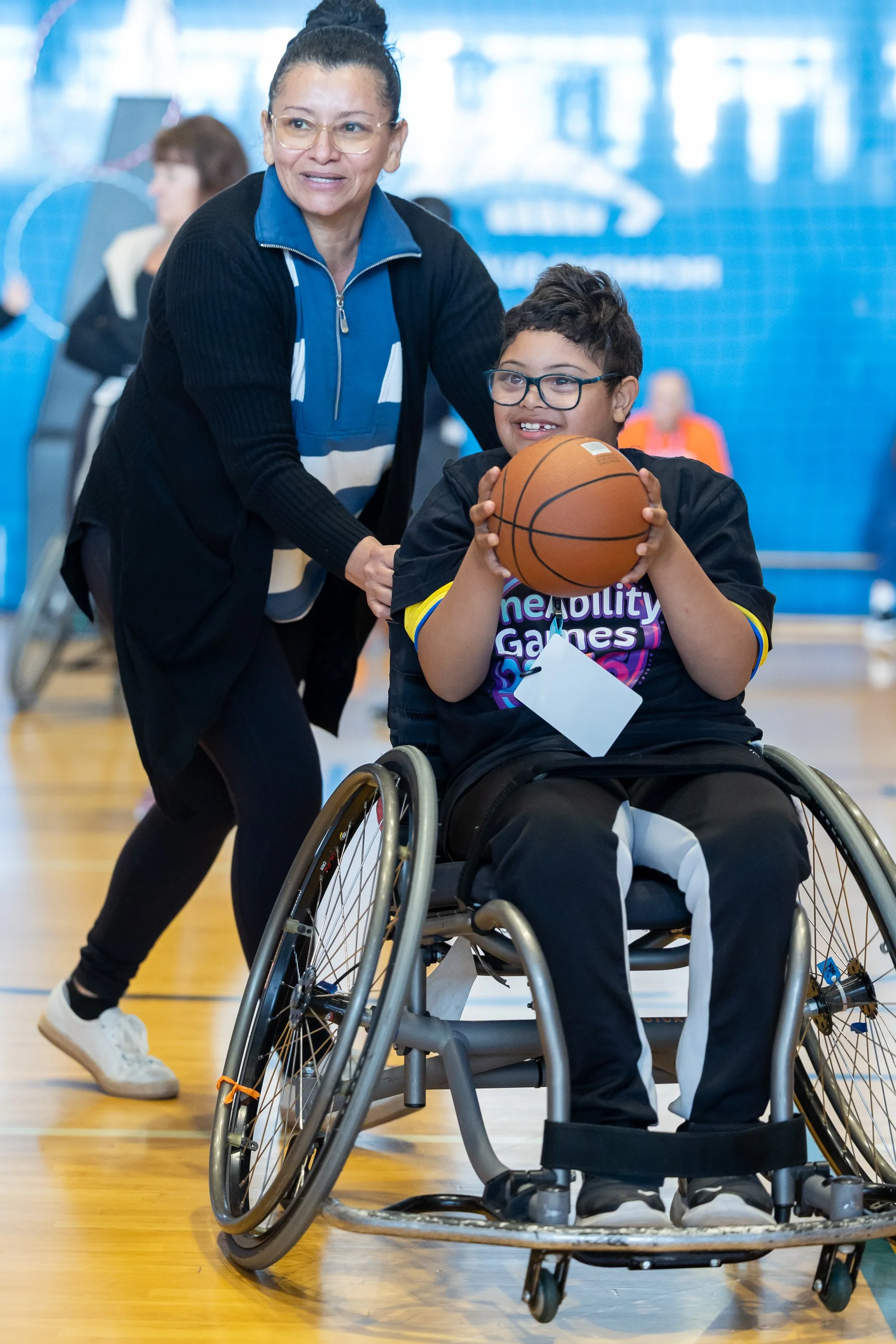 A little boy sits in a sports wheelchair with a big smile on his face, holding a basketball