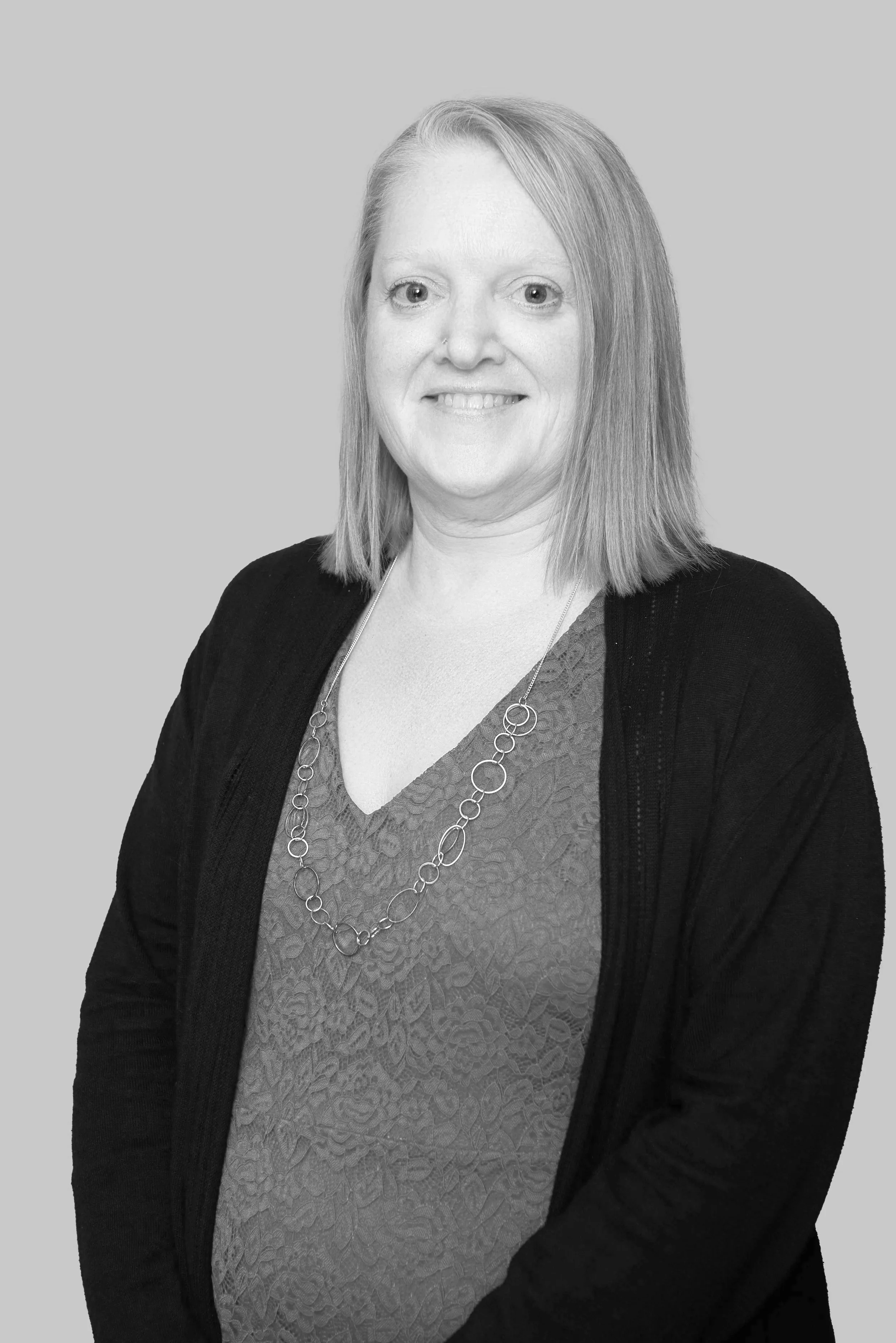 A smiling photo of straight blonde haired Jenn Hobson, a white woman wearing a v neck shirt, black cardigan, and long chain necklace.