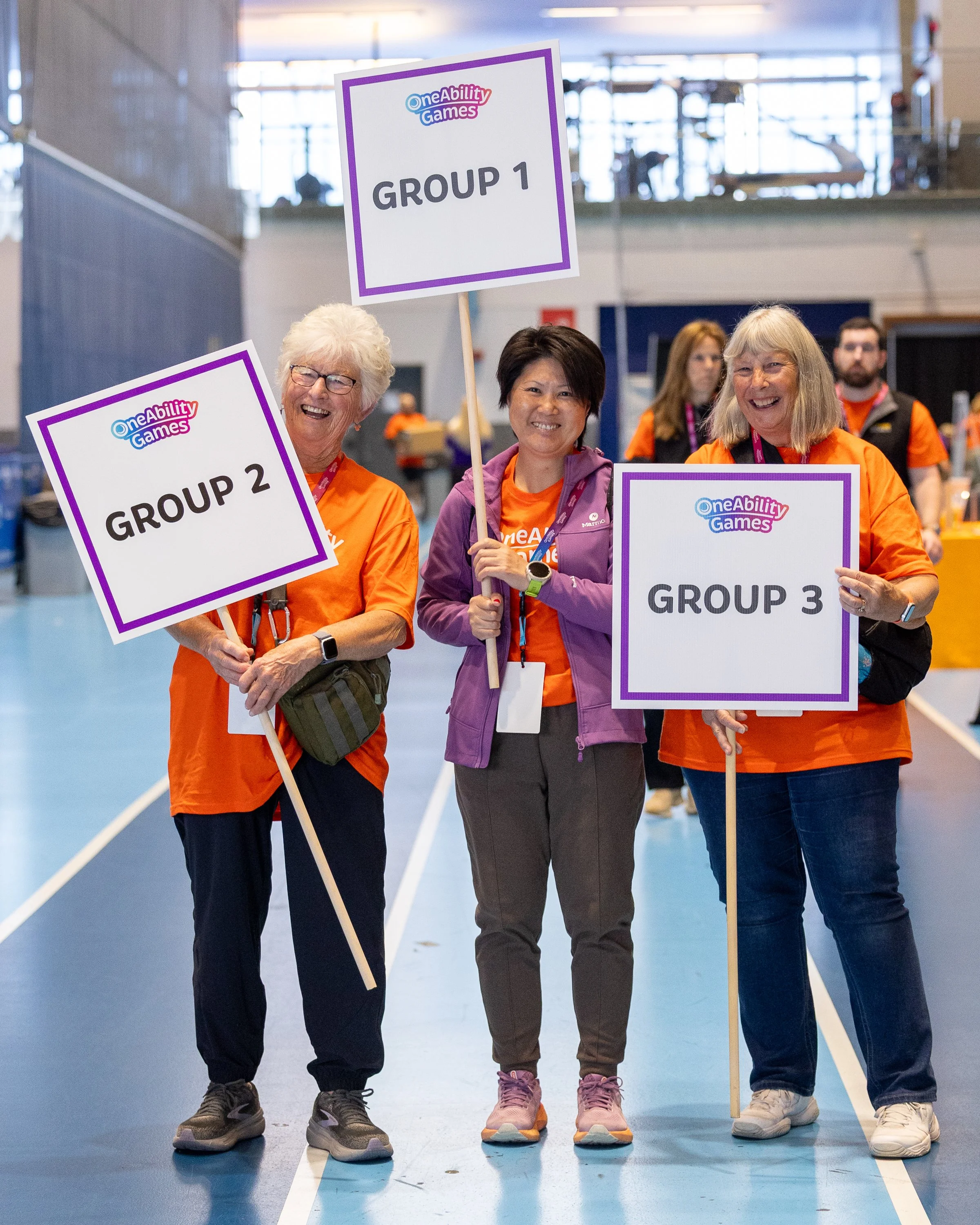 Three women stand side by side, wearing orange volunteer shirts, smiling, and holding group signs