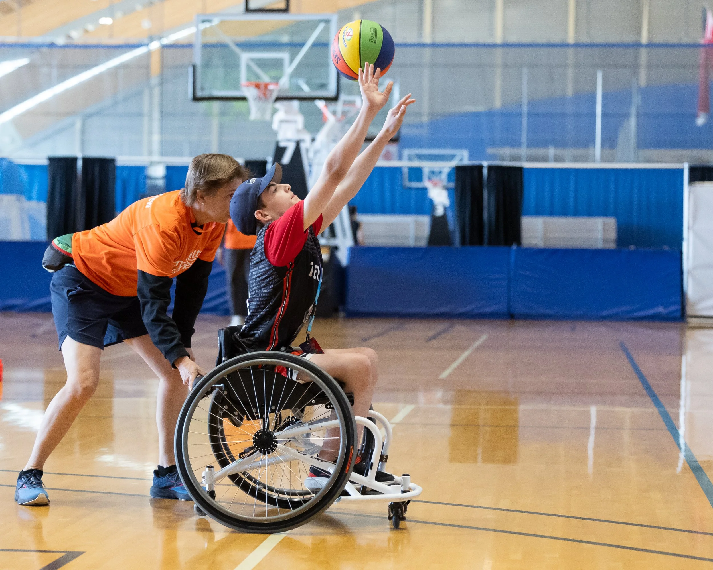 A middle school student sits in a sports wheelchair, arms outstretched, having just thrown a colourful basketball