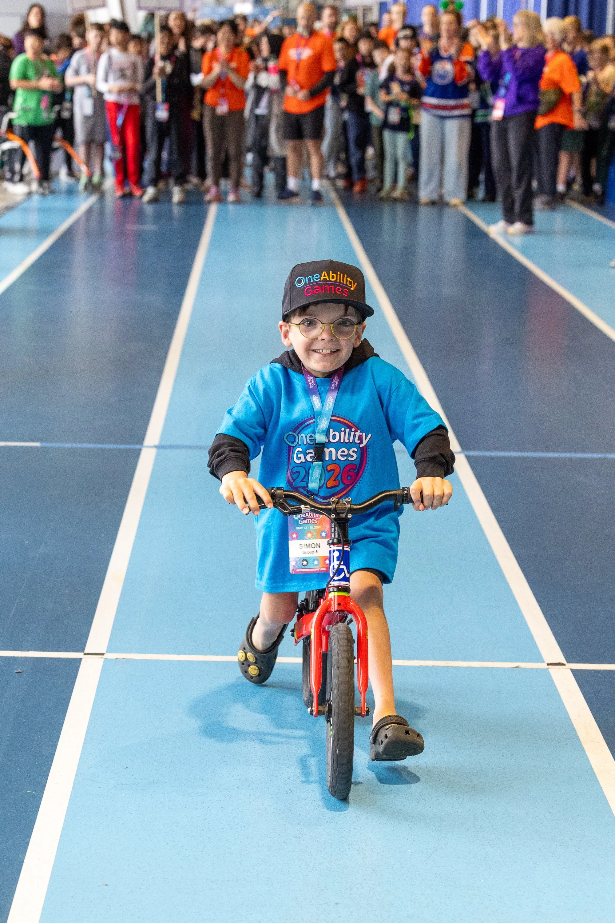 A little boy wearing a blue OneAbility Games shirt rides his bicycle down a track with a crowd of people behind him cheering