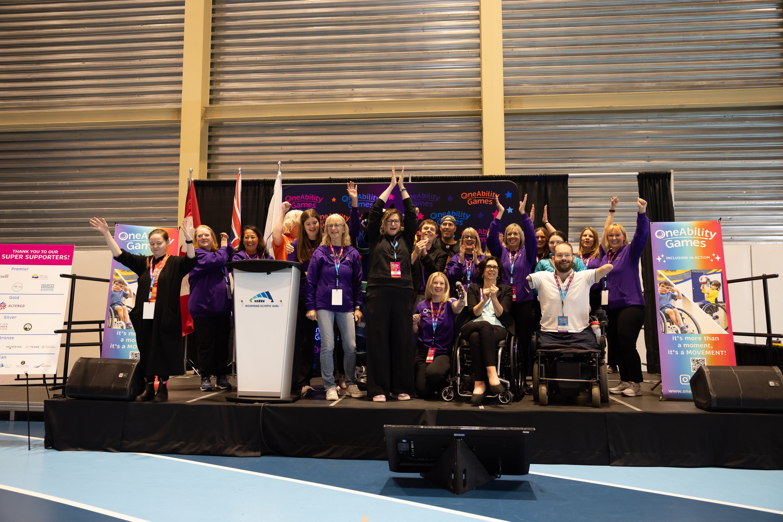 A crowd of people stand and sit in wheelchairs on a stage celebrating the opening of the OneAbility Games