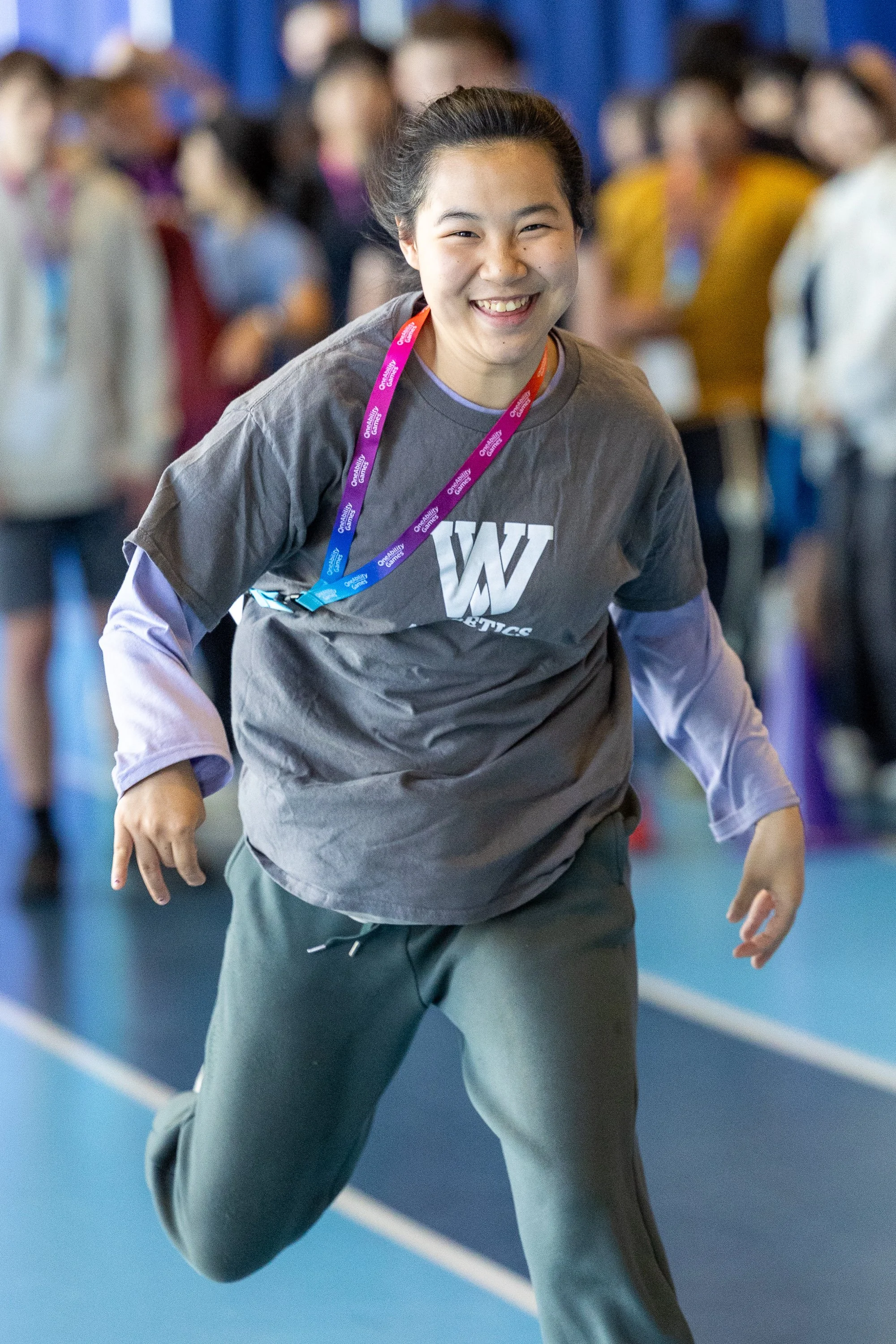 A young girl with black hair, wearing a grey t-shirt runs down a track with a huge smile on her face
