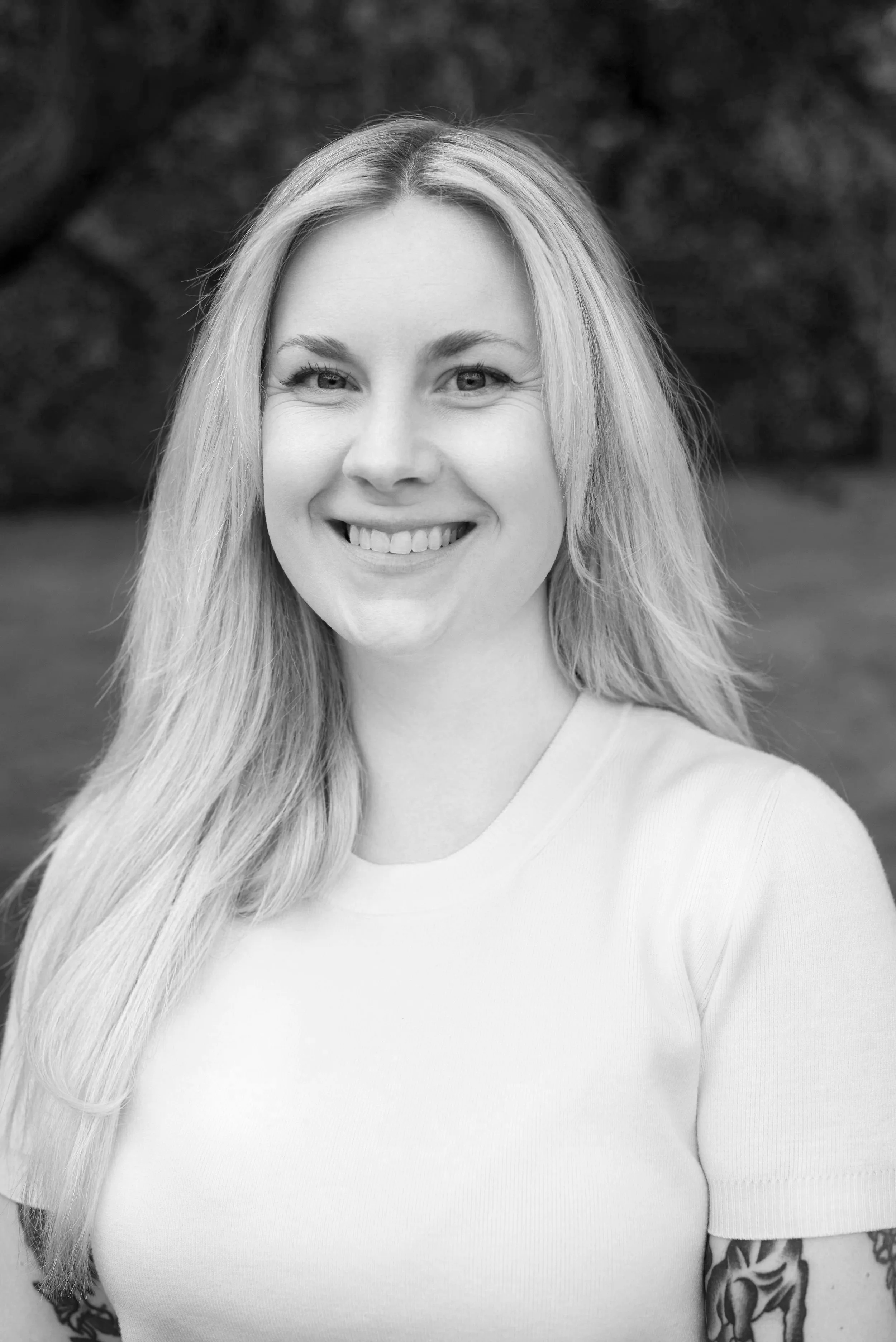 A black and white photo of Leah, a white woman, with long straight blonde hair. She is smiling and wearing a white t-shirt.