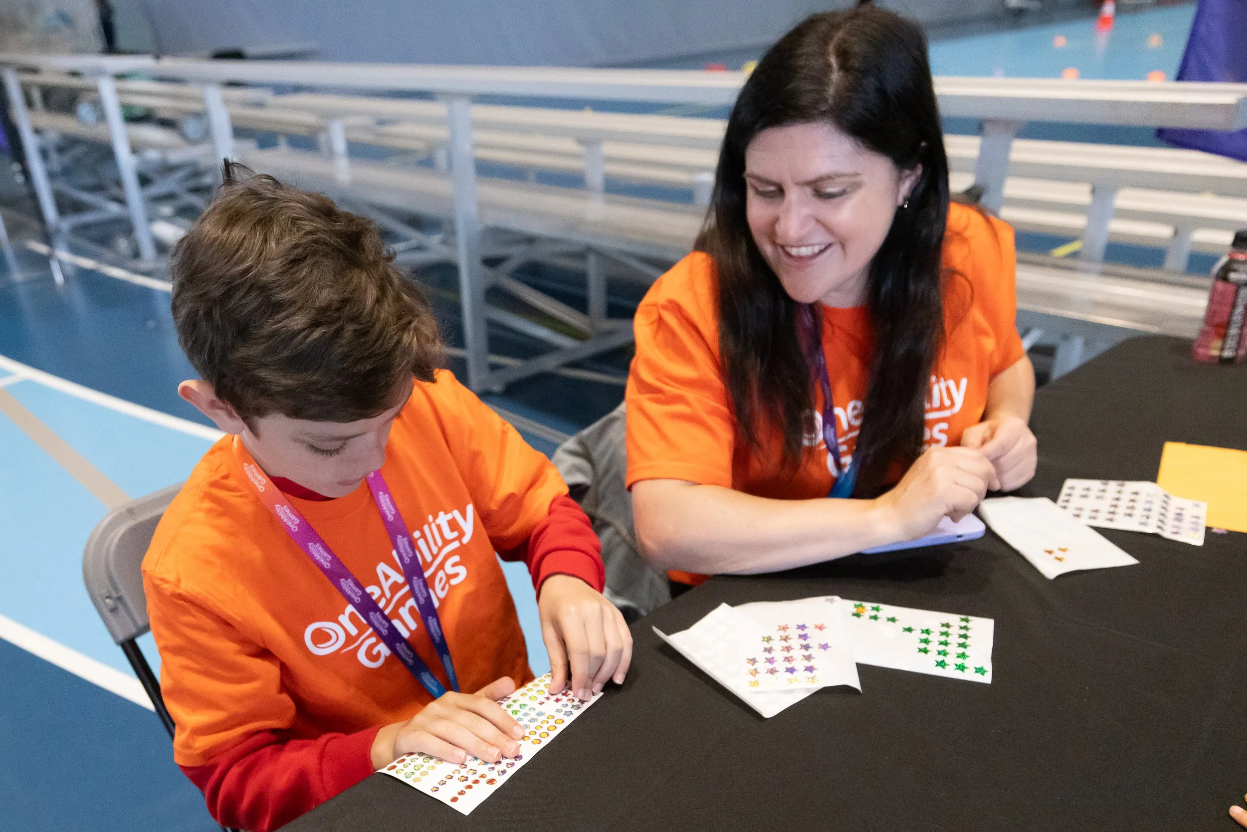 A young boy and his Mom are wearing orange OAG volunteer shirts and peeling stickers