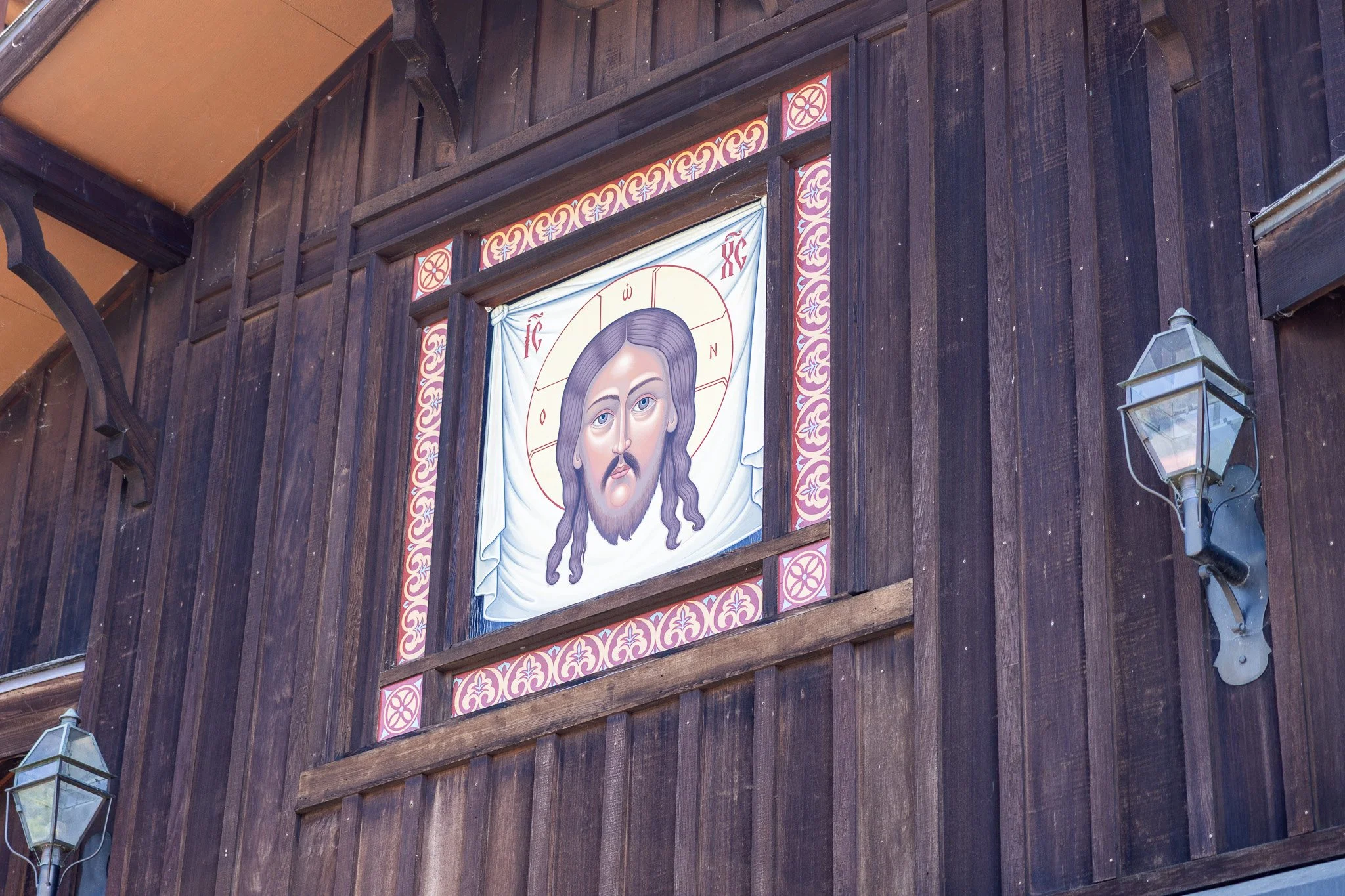 Religious icon of Jesus Christ with long hair, beard, and halo, painted on a cloth backdrop, mounted on a dark wooden wall with decorative border, held by wooden support beams, with two lantern-style outdoor lights on each side.