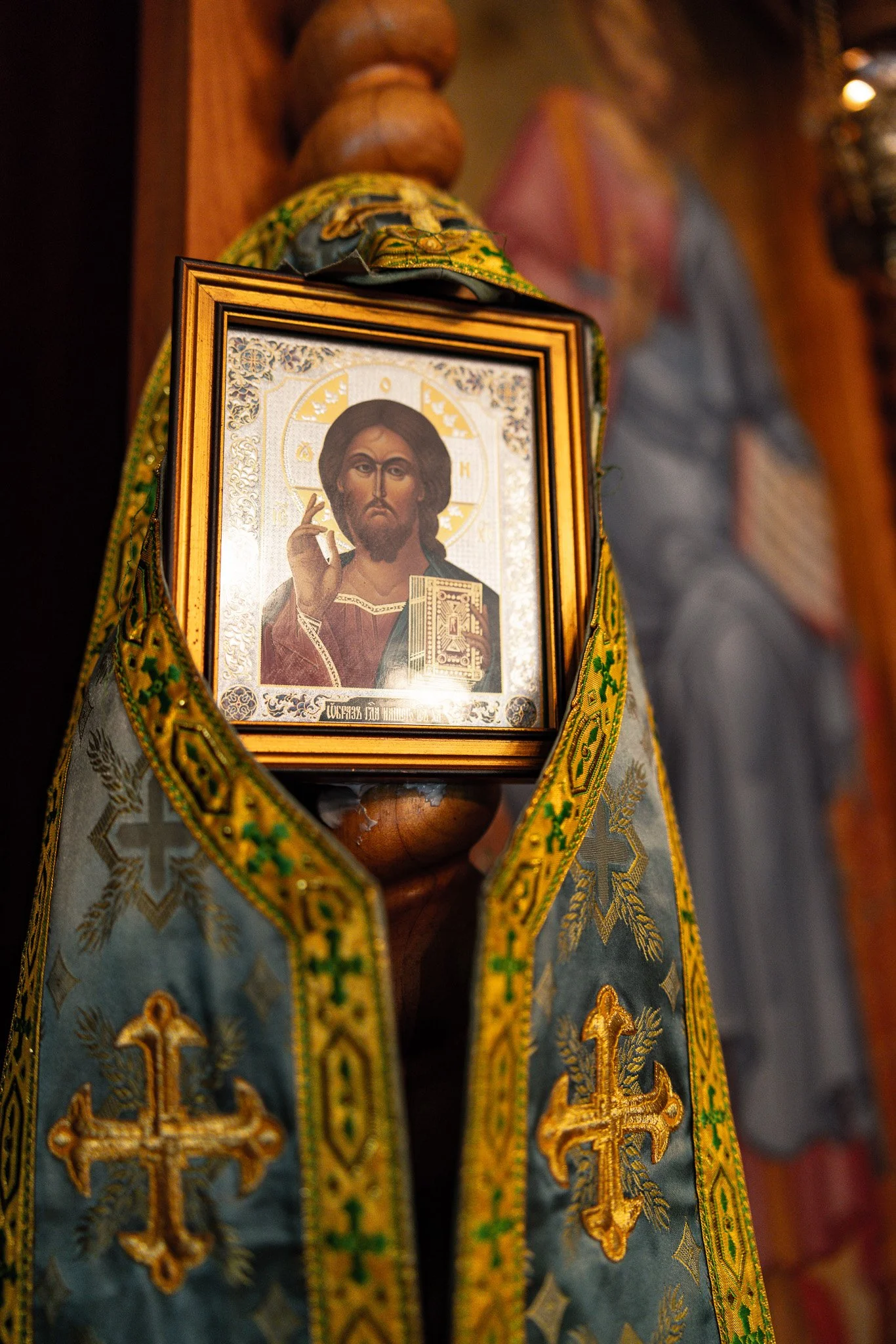 An icon of Jesus Christ with a golden halo and hand raised in blessing, framed and draped with a blue and gold embroidered cloth with crosses, inside a church setting.