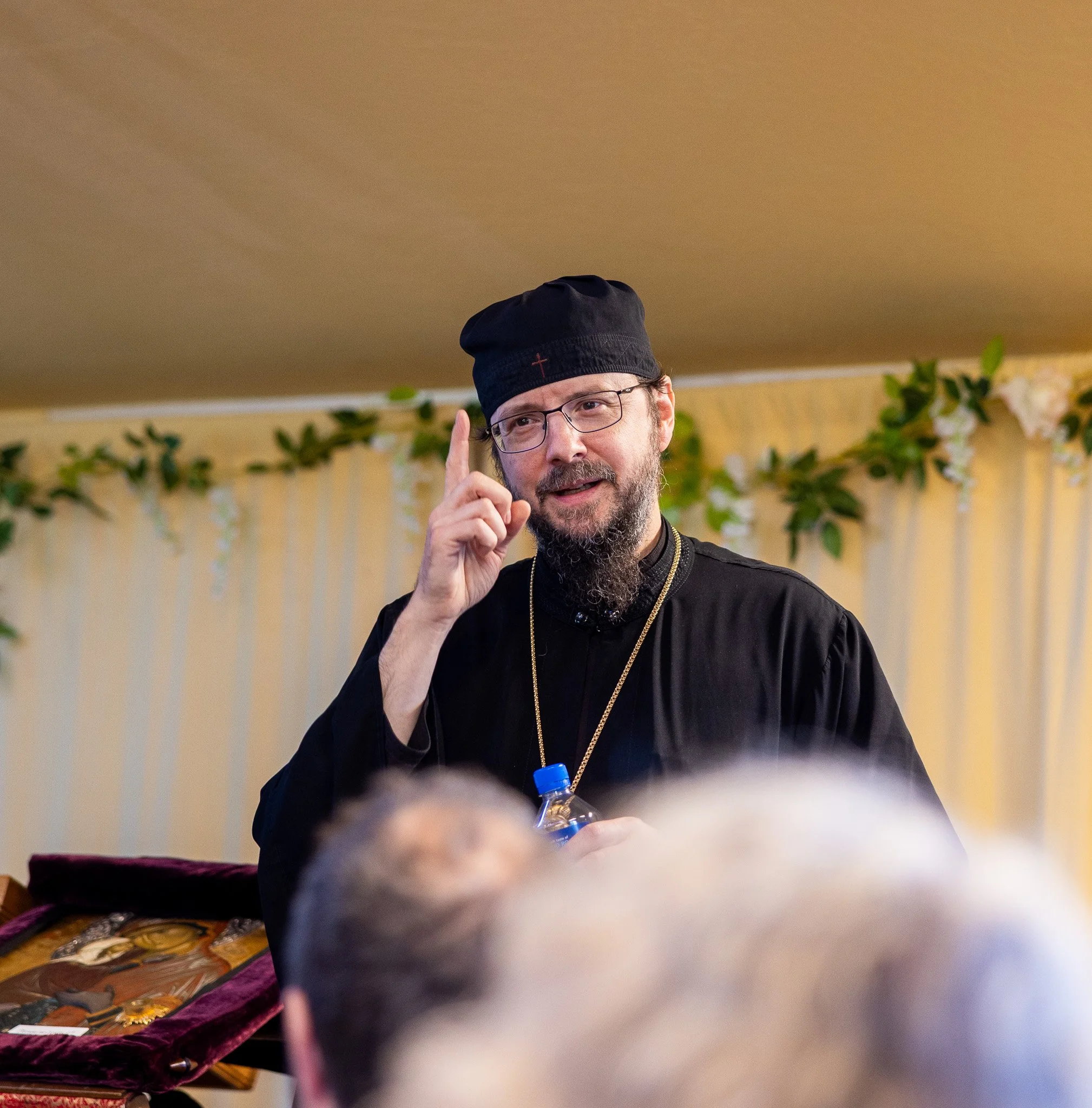 Abbot Sergius of St. Tikhon's Seminary wearing glasses, a black robe, and a black cap, holding a water bottle and raising one finger while speaking to a group. The background has yellow curtains and greenery.
