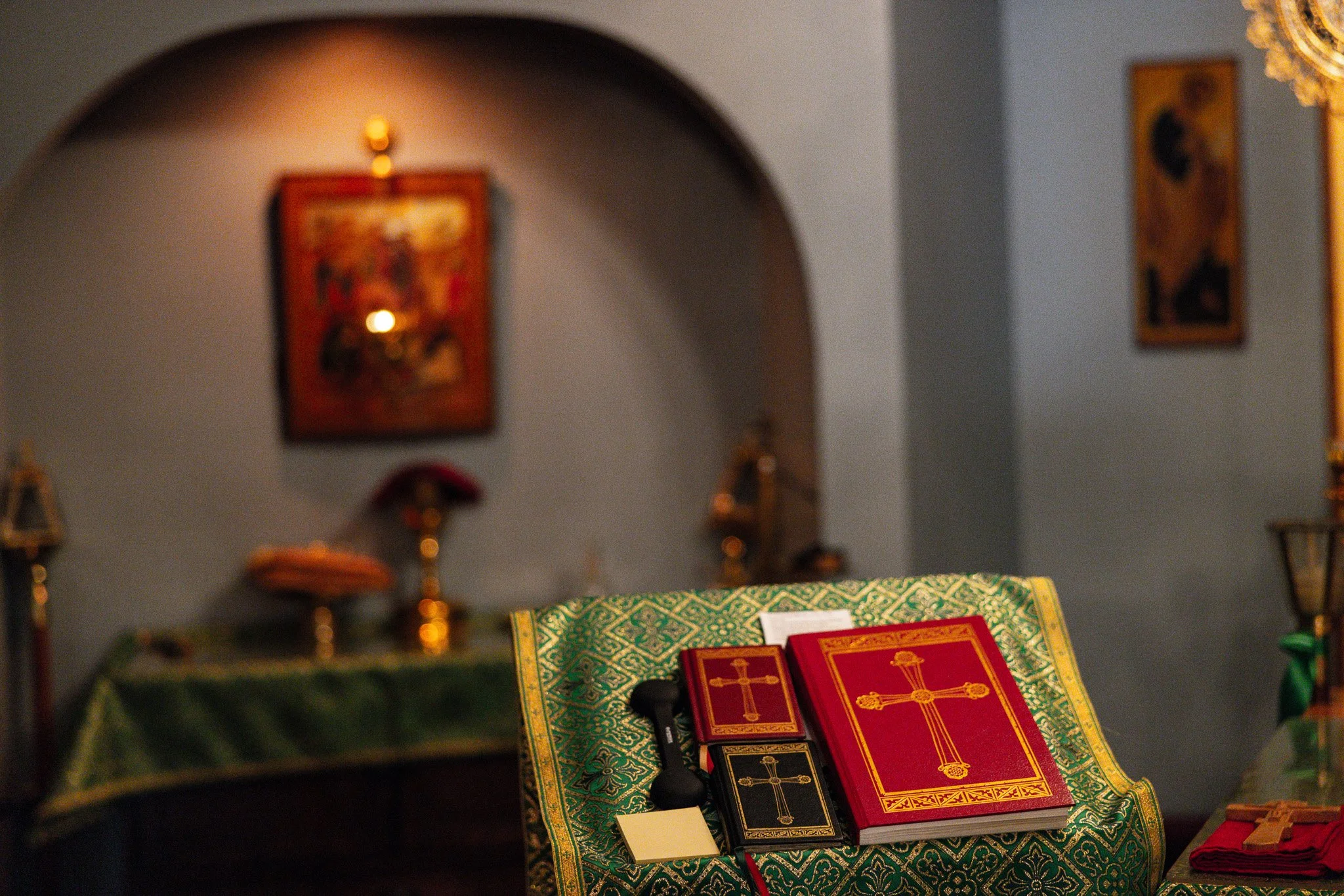 orthodox altar with red and black prayer books featuring gold crosses, a microphone, and papers, set on a green cloth with gold patterns, inside a church with icons and religious artwork in the background.