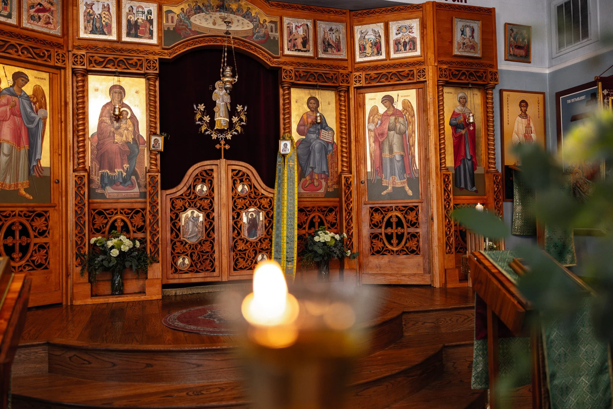 Inside an Orthodox church, an ornate wooden iconostasis with religious icons and paintings, lit by candles, with flowers and religious objects placed in front.