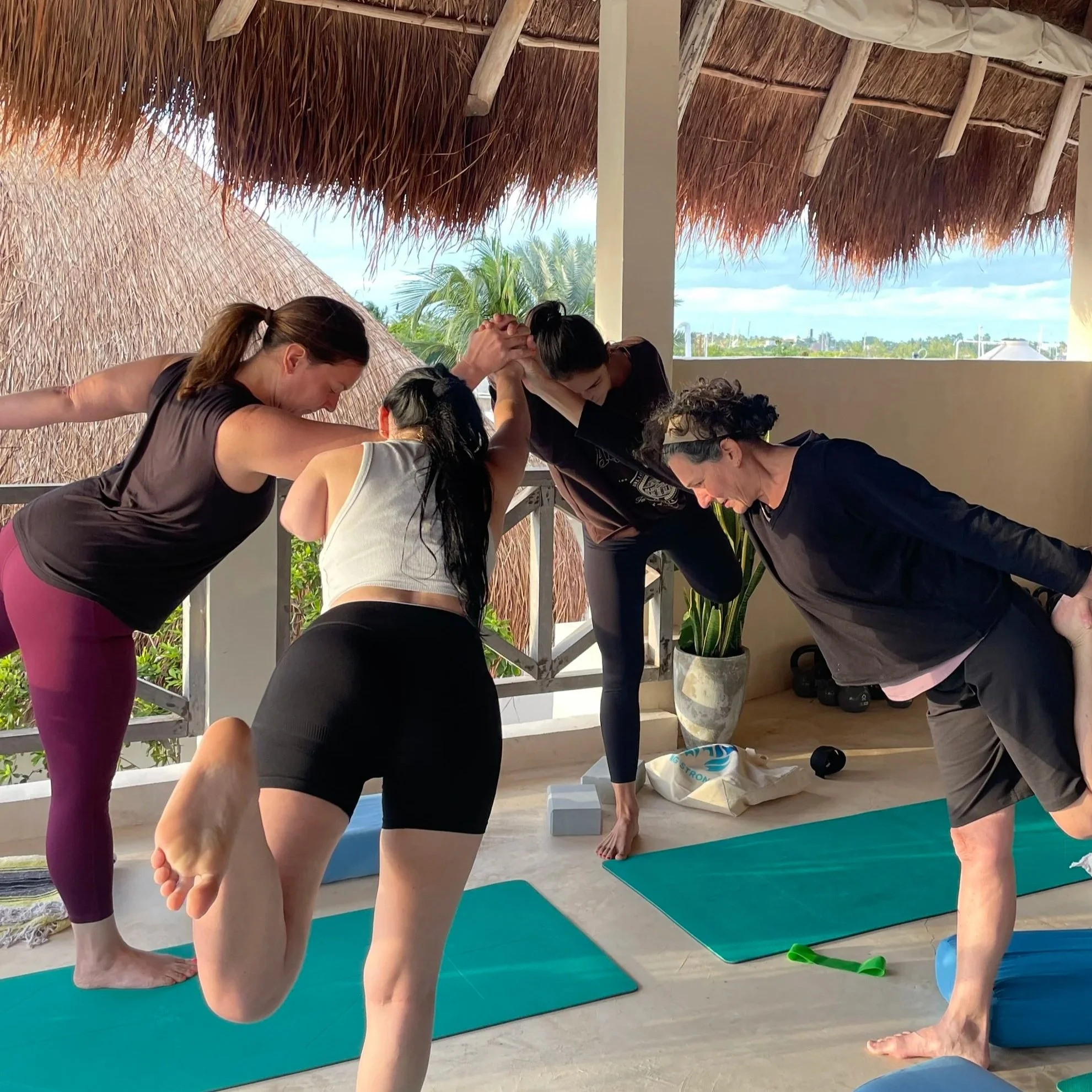 Group of four women practicing yoga on mats on a covered outdoor patio with palm trees and blue sky in the background.
