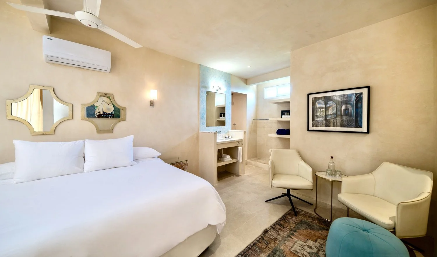 A hotel room with a bed, two chairs, a small table, and a bathroom area with a sink and mirror.