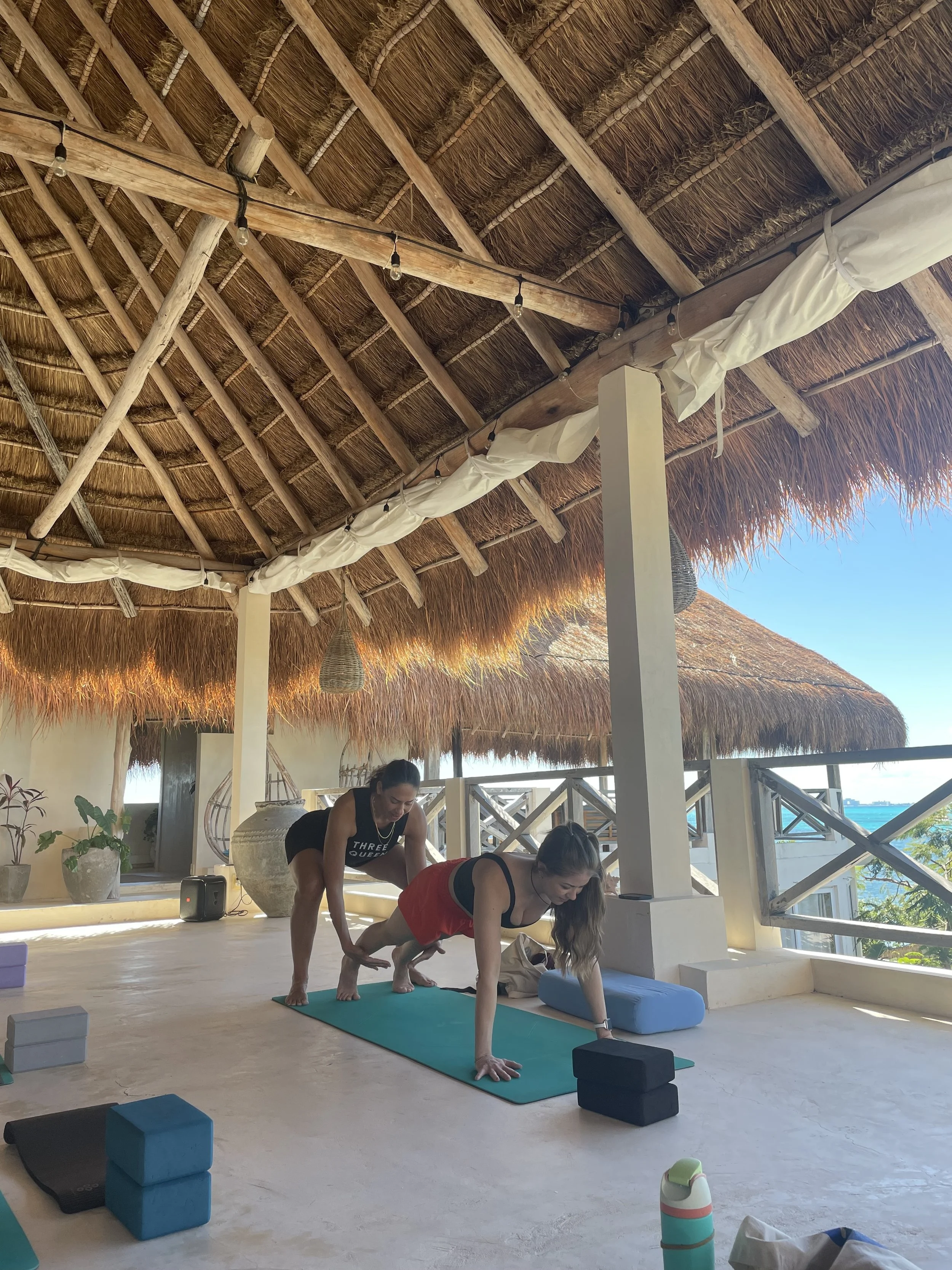 Two women practicing yoga on mats inside a thatched-roof open-air space, with natural light and ocean view in the background.