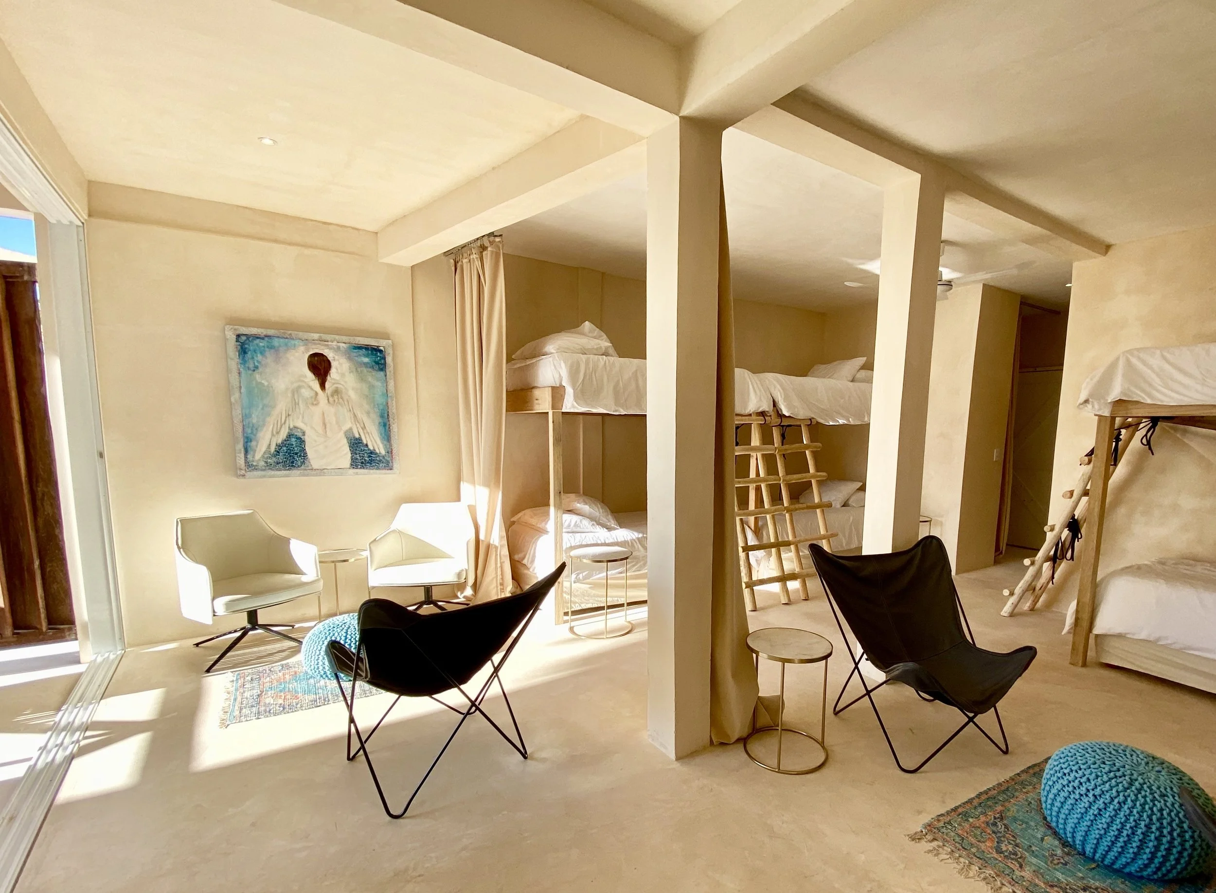 Interior of a room with beige walls and ceiling, featuring a sitting area with two white chairs, black butterfly chair, and small round tables. There are floor-to-ceiling bunk beds with wooden ladders on each side. Artwork of an angel with wings and a red heart is hung on the wall. Natural light is entering through sliding glass doors on the left, with a colorful rug and a blue crocheted pouf on the floor.