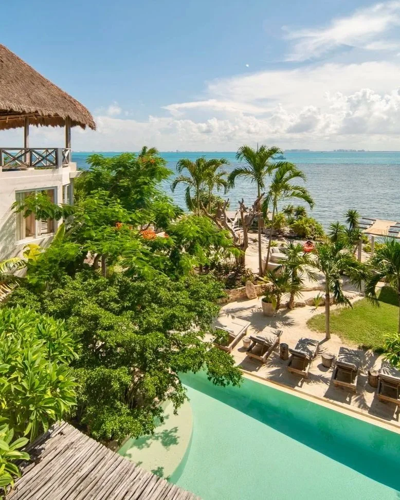 A tropical seaside resort with a view of the ocean, palm trees, lush greenery, a pool, sun loungers, and a thatched-roof building.