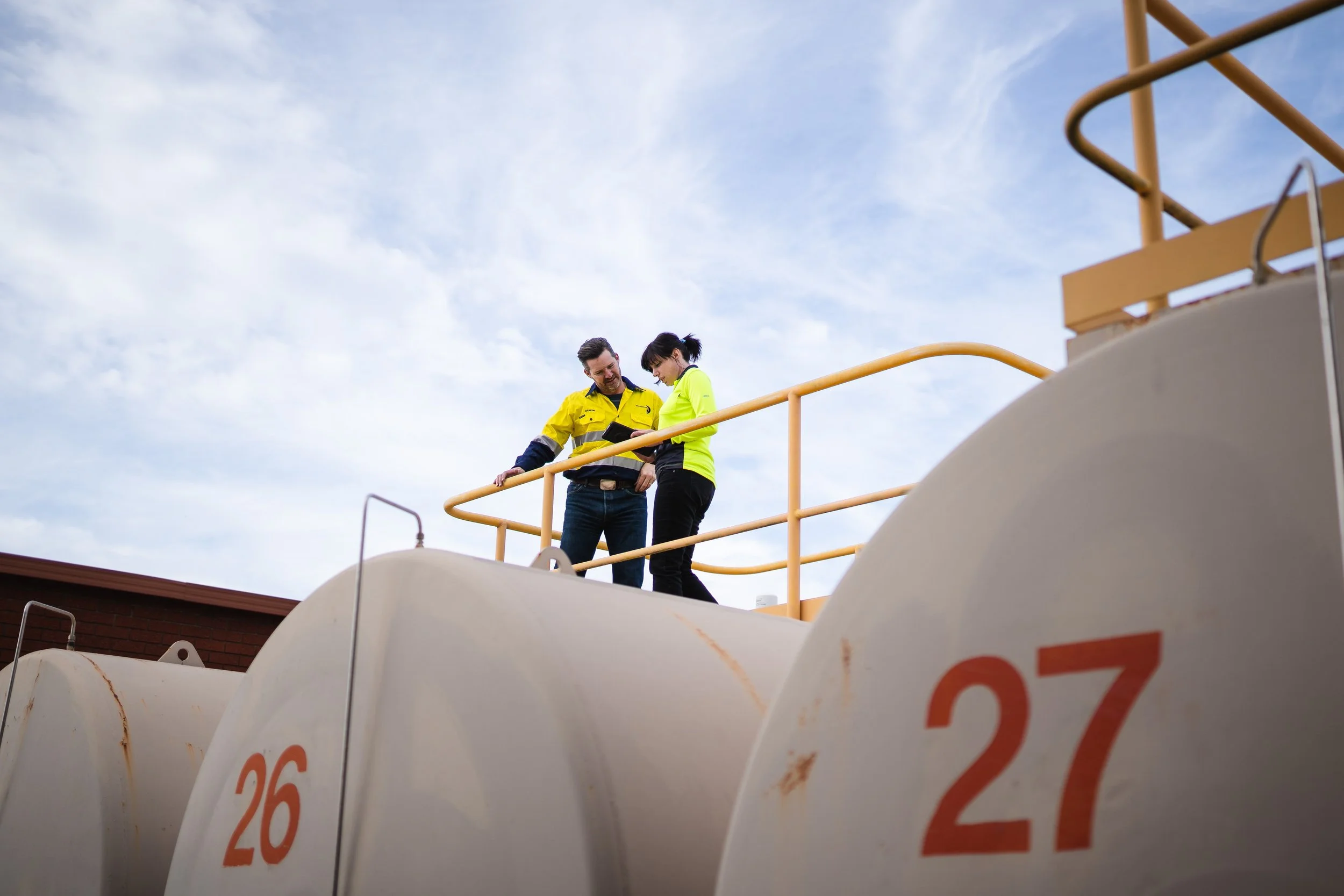 Two workers in safety gear stand on a platform above large industrial tanks numbered 26 and 27.