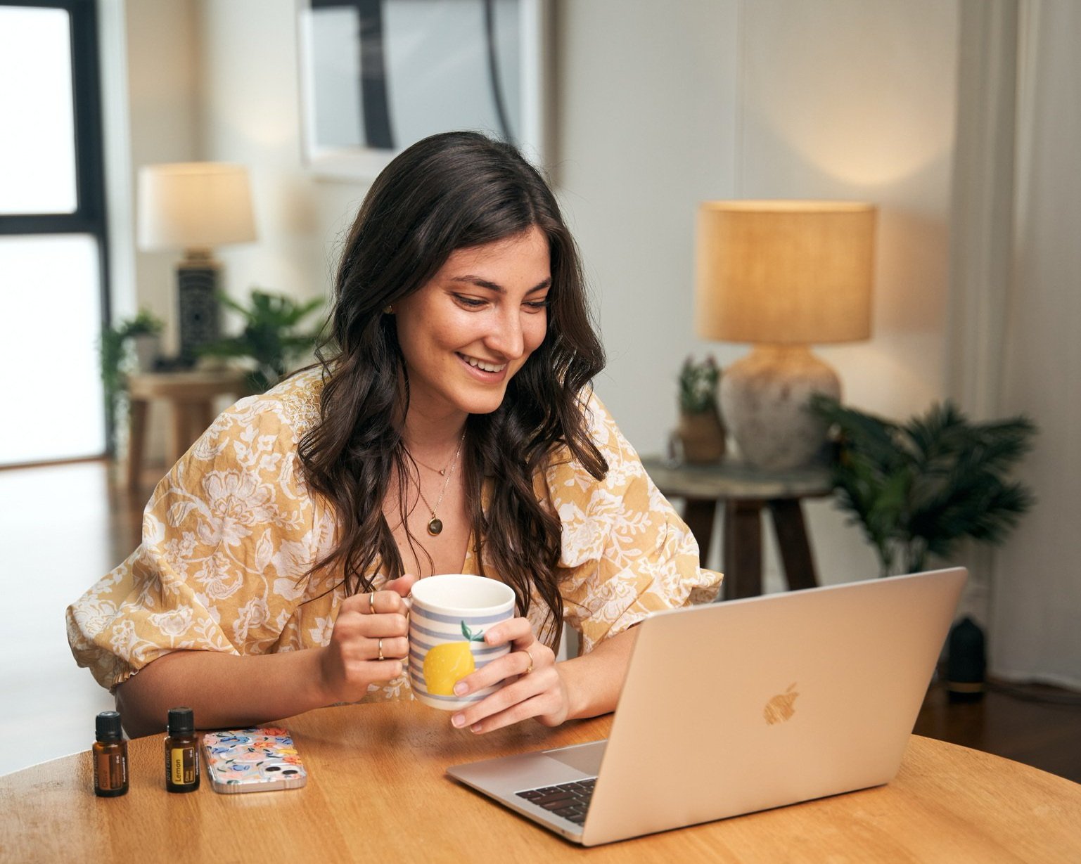 Woman smiling at a laptop while holding a coffee mug in a cozy room with plants and lamps.
