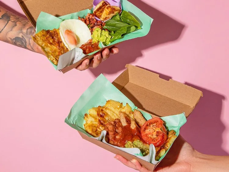 Two hands holding cardboard food containers with assorted meals, including items like eggs, spinach, guacamole, hash browns, tomatoes, and sauce, against a pink background.