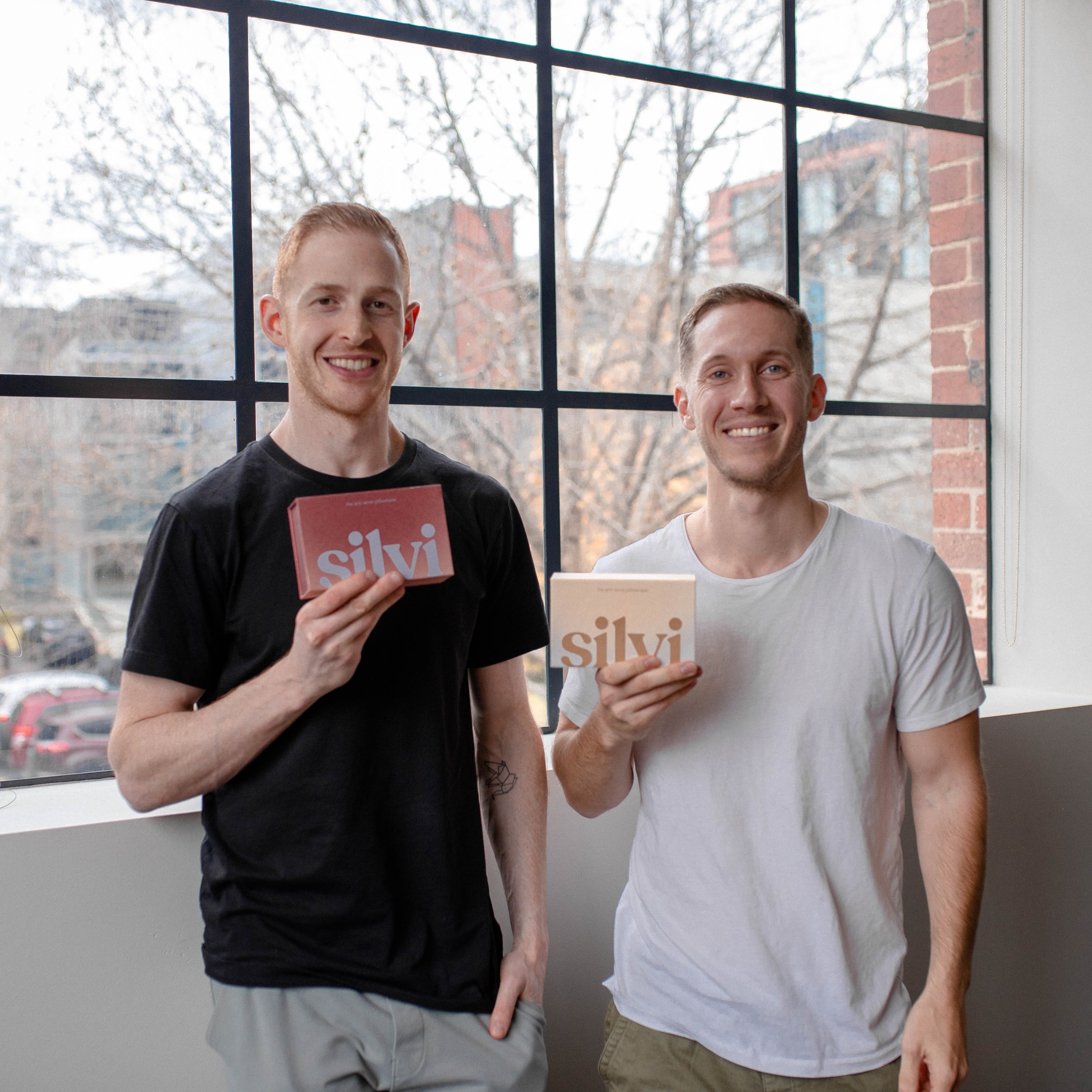Two men smiling and holding Silvi branded boxes in an indoor setting with large windows in the background.