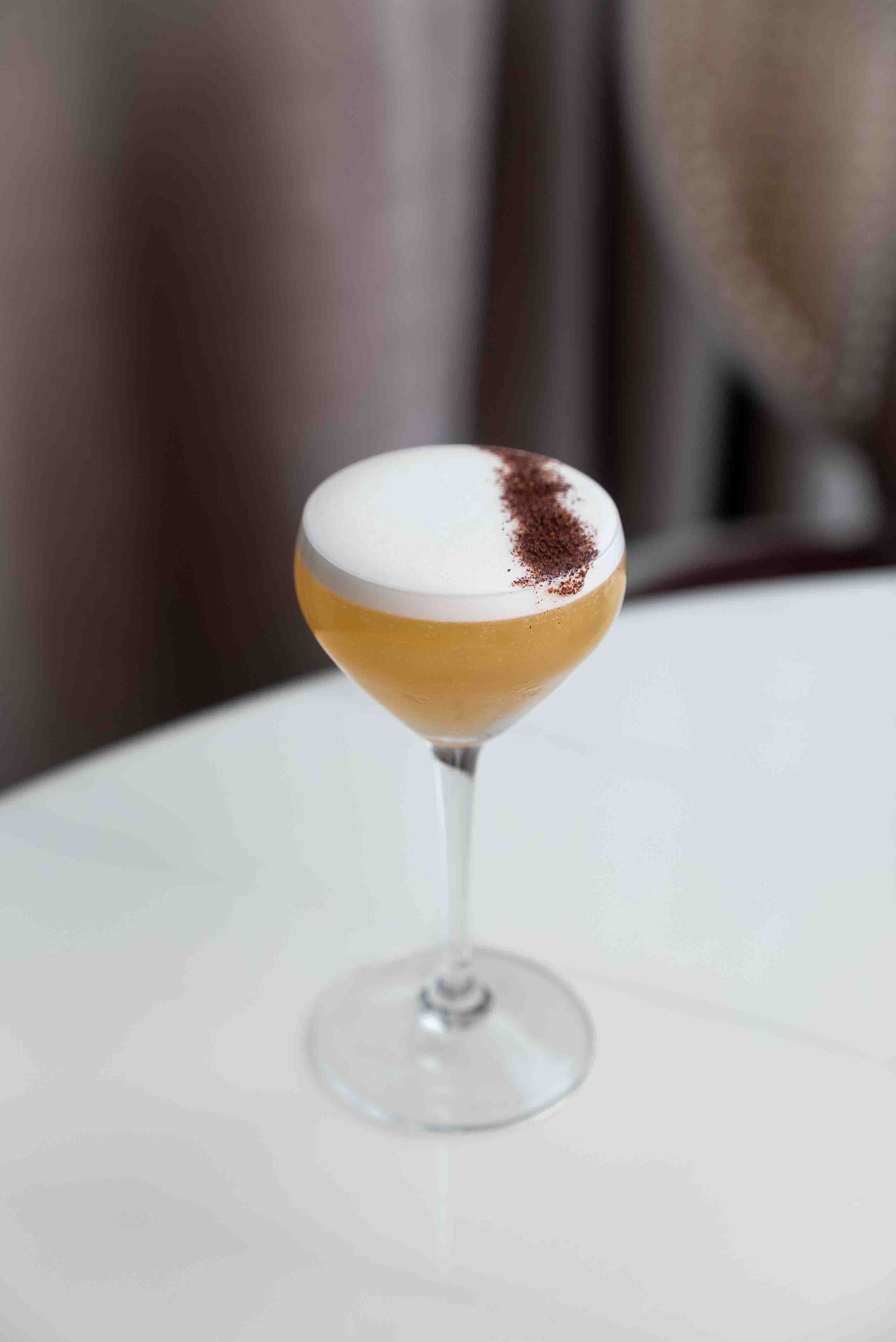 Cocktail in a coupe glass with frothy top and cocoa garnish on a table.
