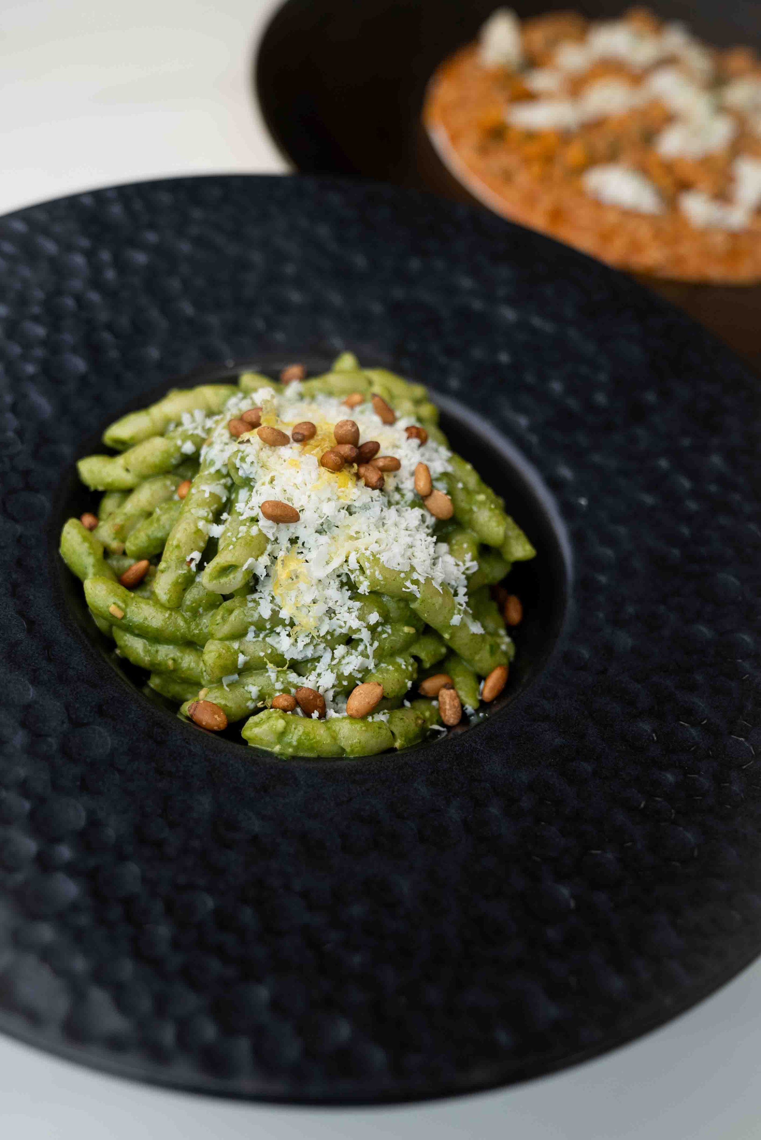 A dish of pasta with green pesto sauce, garnished with grated cheese and pine nuts, served on a black plate.
