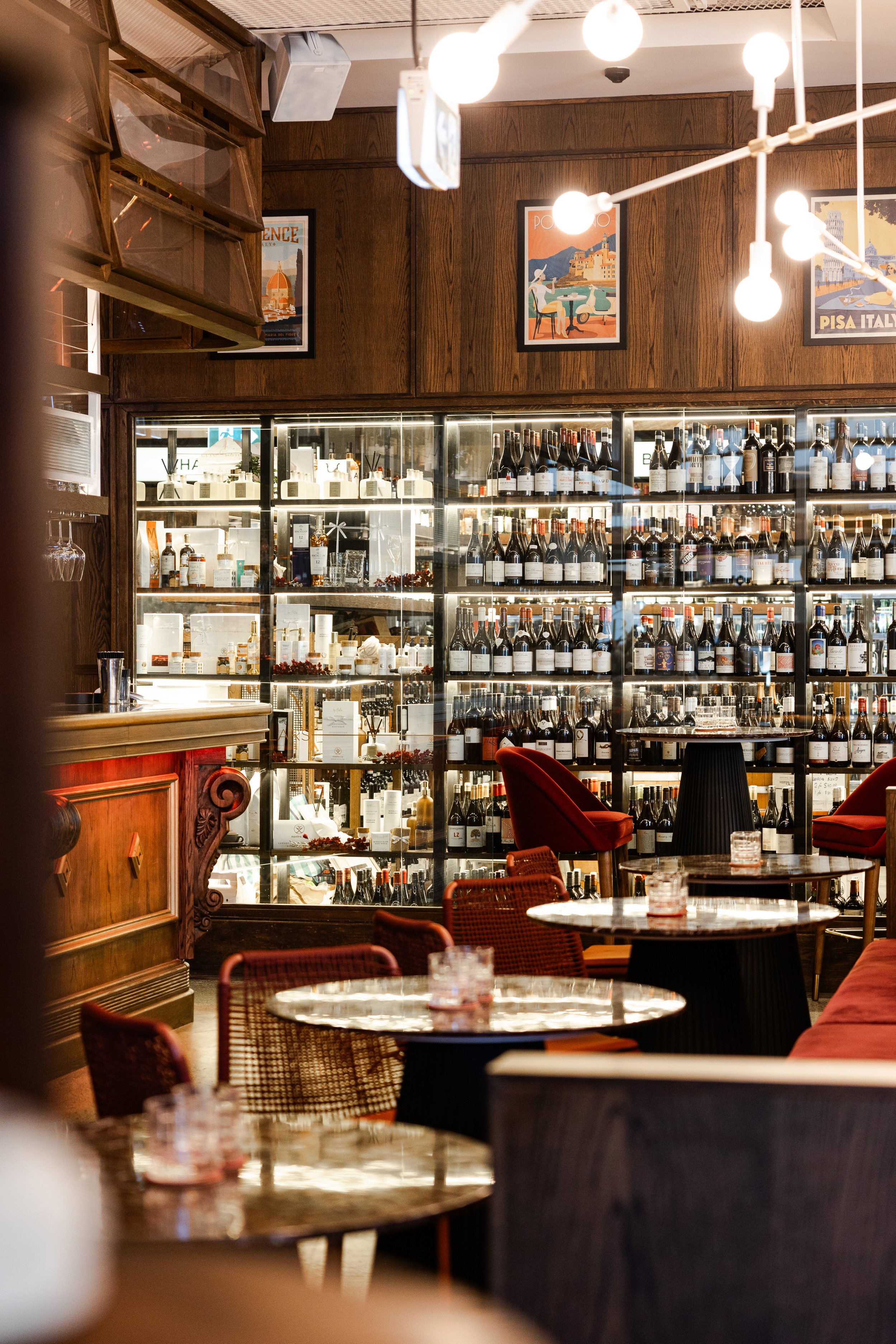 Interior of a cozy wine bar with wooden decor, round tables, red velvet chairs, and a large display of wine bottles on shelves. Stylish lighting hangs from the ceiling.