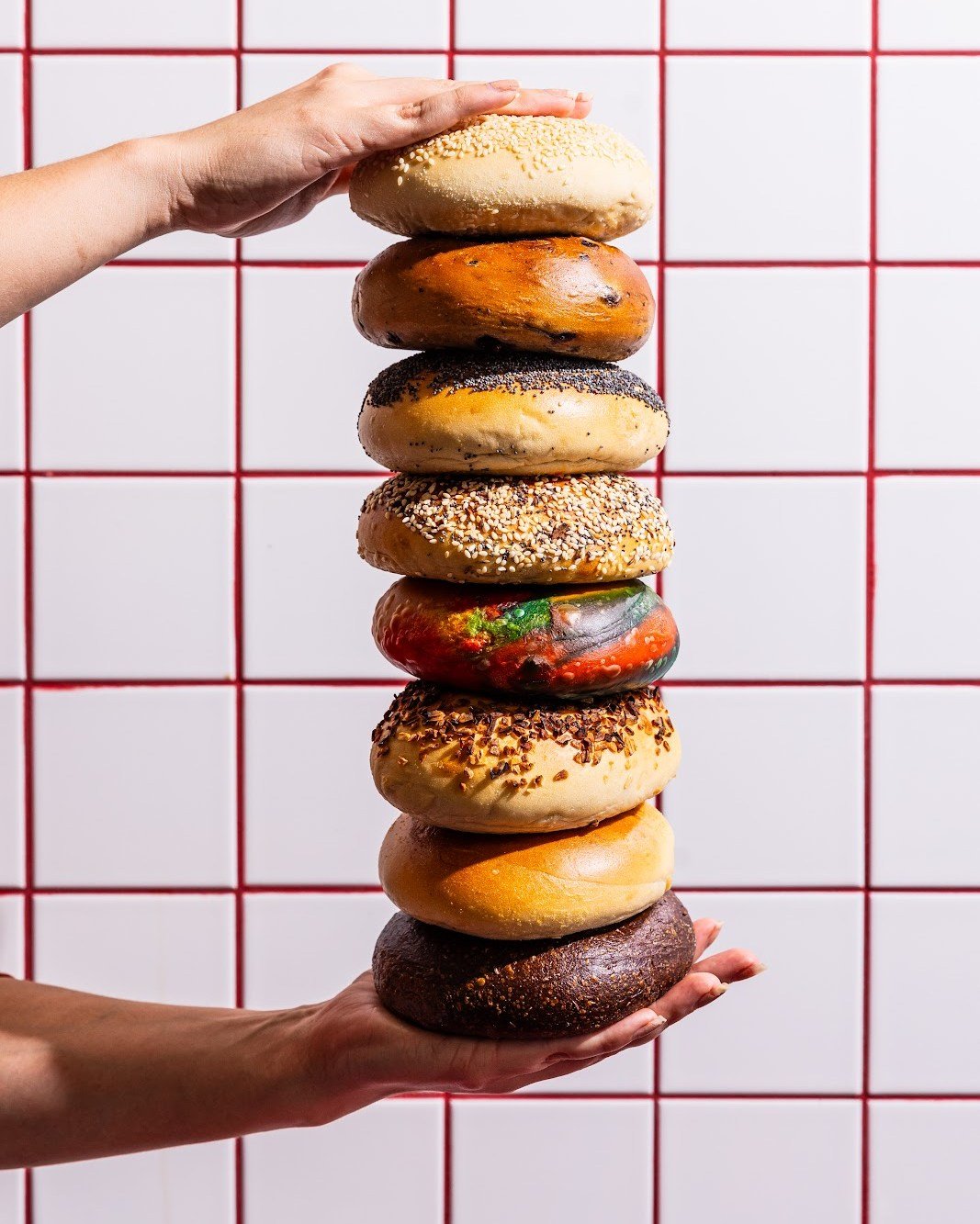 Stack of assorted bagels held by two hands in front of a tiled wall.