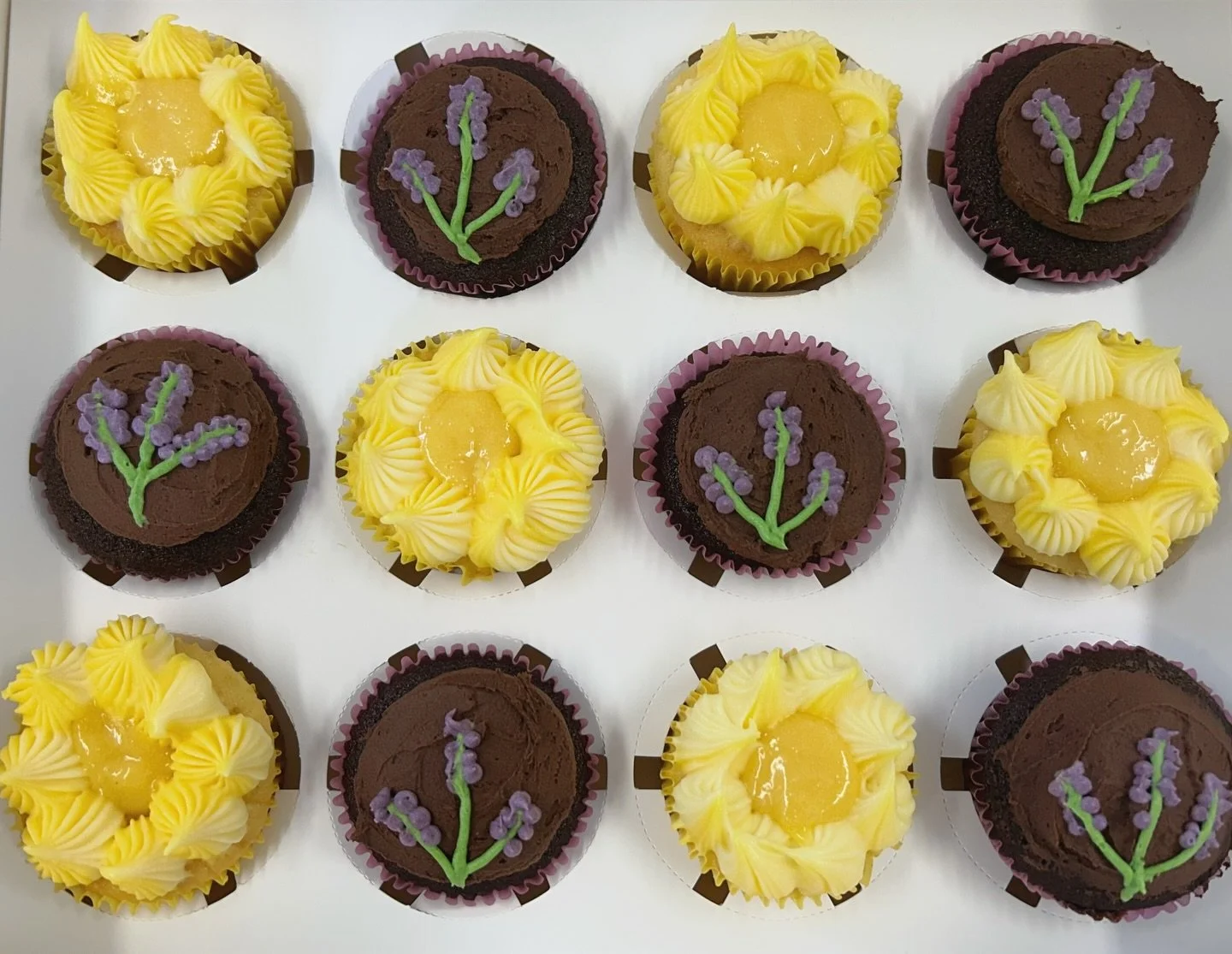 A tray of twelve cupcakes featuring yellow and chocolate frosting, with some decorated with lavender-colored flower designs and others with a yellow glaze topping.