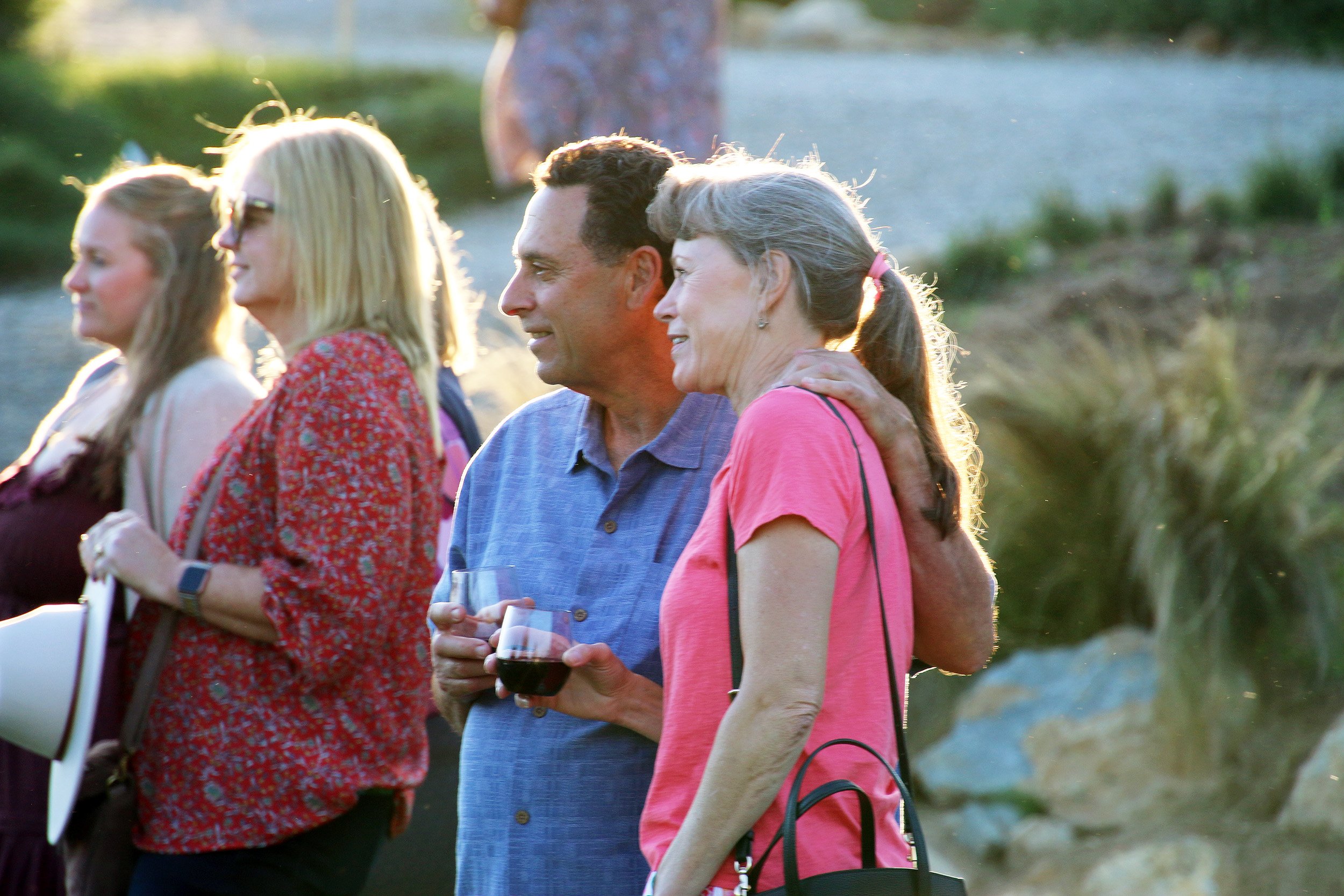 Group of people outdoors at an event, two with drinks, embracing and talking