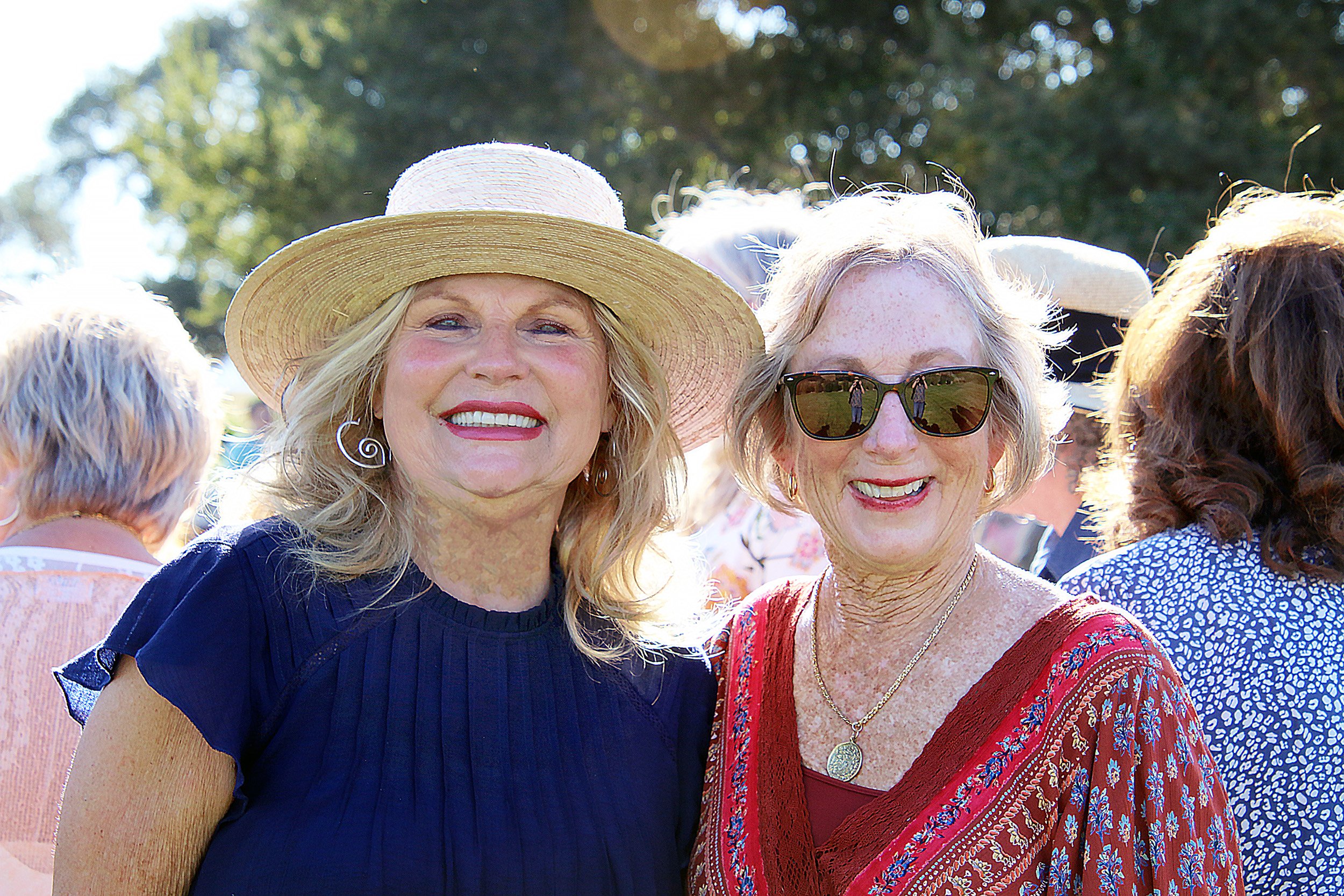 Two women smiling at an outdoor event, one wearing a straw hat and the other sunglasses.