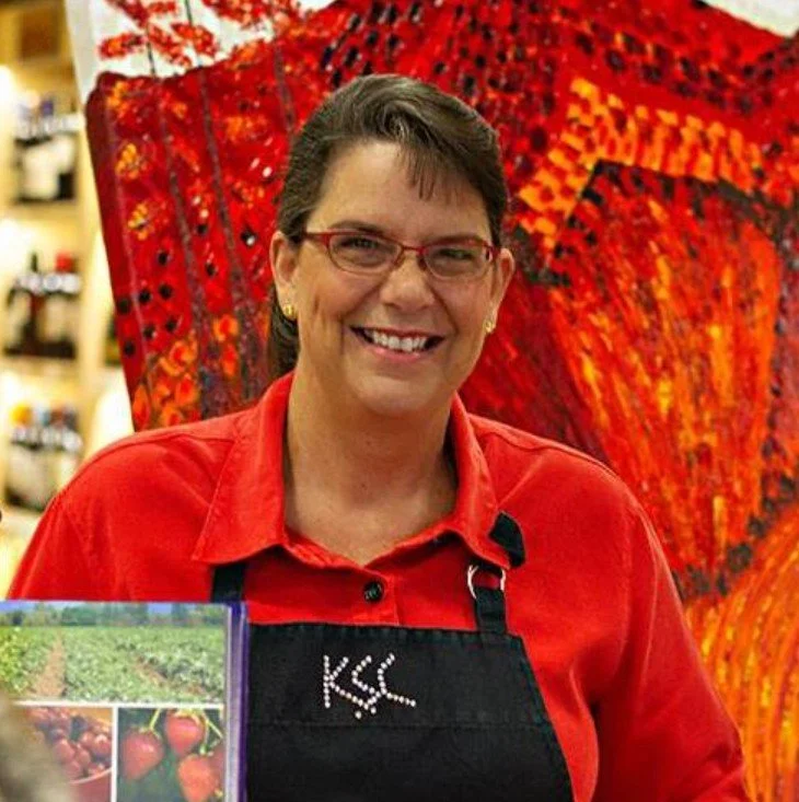A woman wearing glasses, a red shirt, and an apron, smiling in front of a large painting with red, orange, and black patterns.