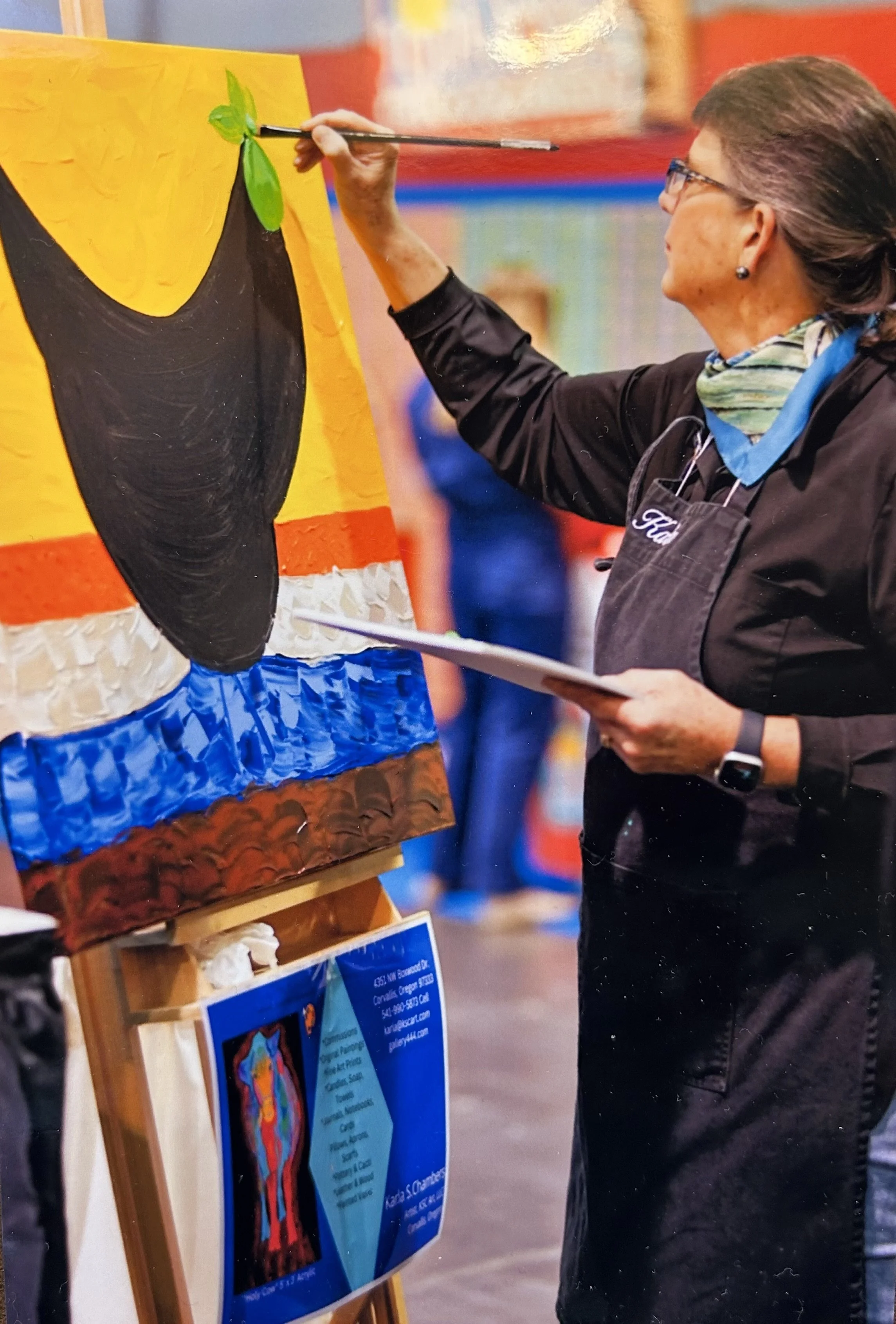 A woman painting a colorful abstract art piece on a canvas, holding a paintbrush and a palette at an indoor art event.