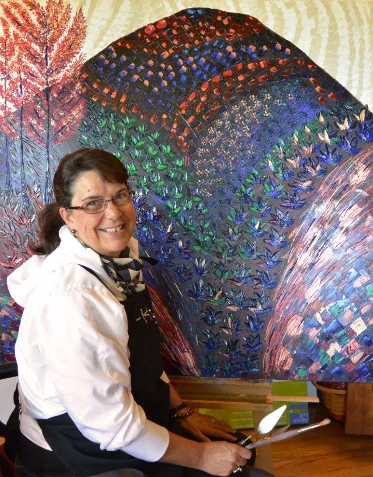 A woman smiling and sitting in front of a vibrant, colorful abstract painting featuring a rainbow of colors with layered patterns and textures.