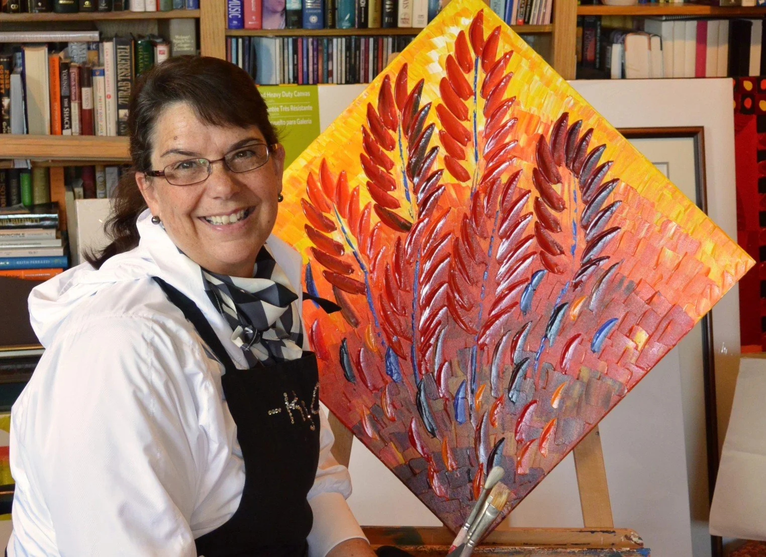 A smiling woman with glasses, wearing a white shirt, black apron, sitting next to a vibrant floral painting with orange, red, yellow, blue, and purple brushstrokes on a diamond-shaped canvas in an art studio with bookshelves in the background.