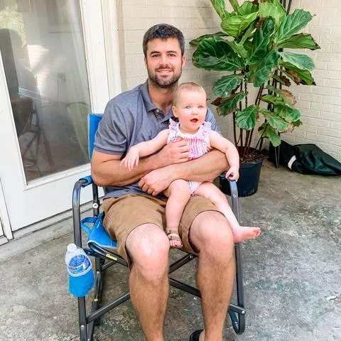 A man with a beard sitting outdoors on a blue folding chair, holding a smiling baby girl in his lap. They are near a white door with glass, and behind them is a large potted green plant and some outdoor items.