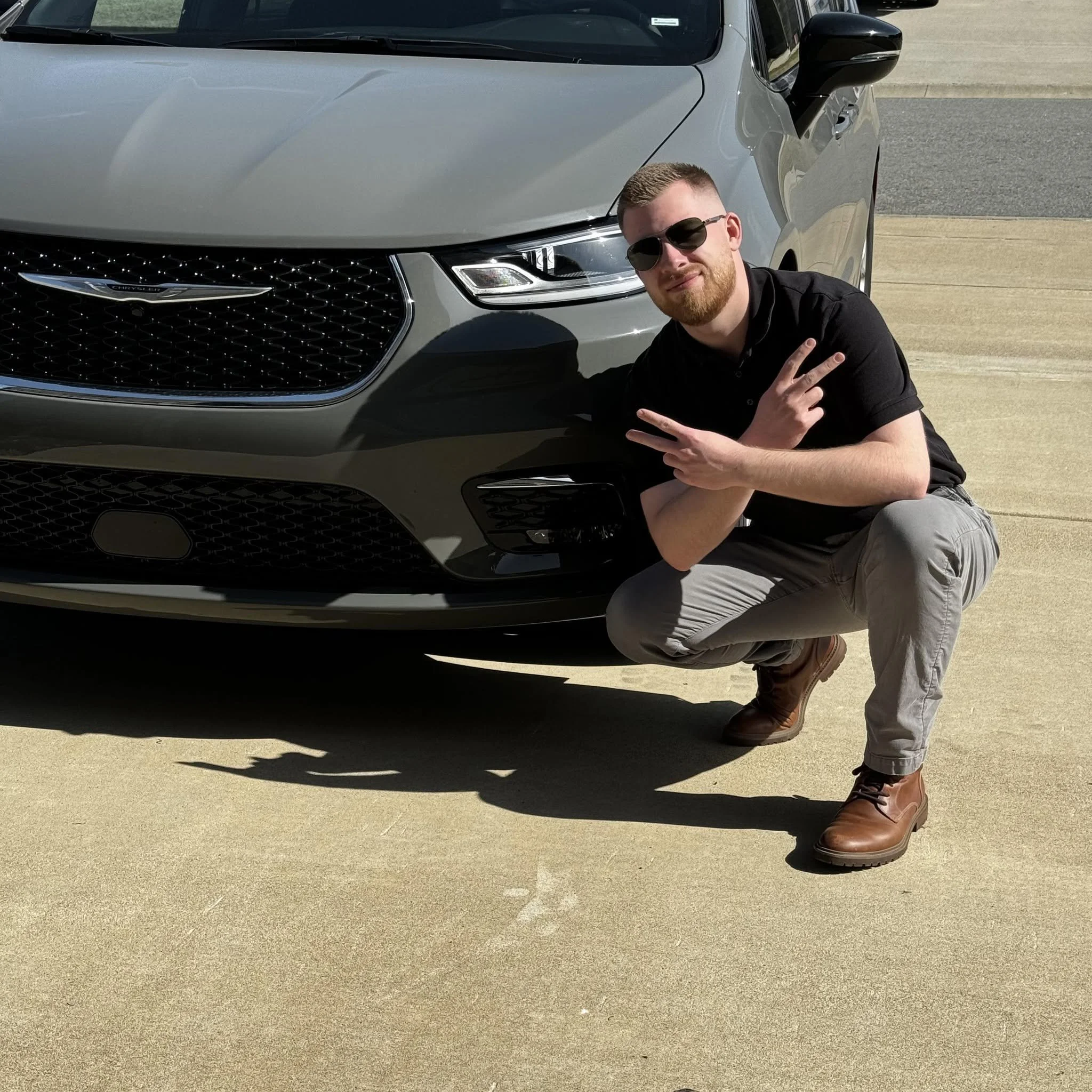 A man with sunglasses and a beard making a peace sign kneels next to a gray Chrysler vehicle in a parking lot.