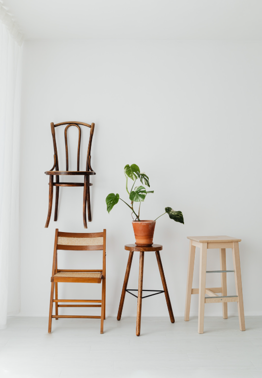 Minimalist interior with a wooden chair, small table with a potted plant, and a stool against a white wall. Another wooden chair is mounted on the wall.