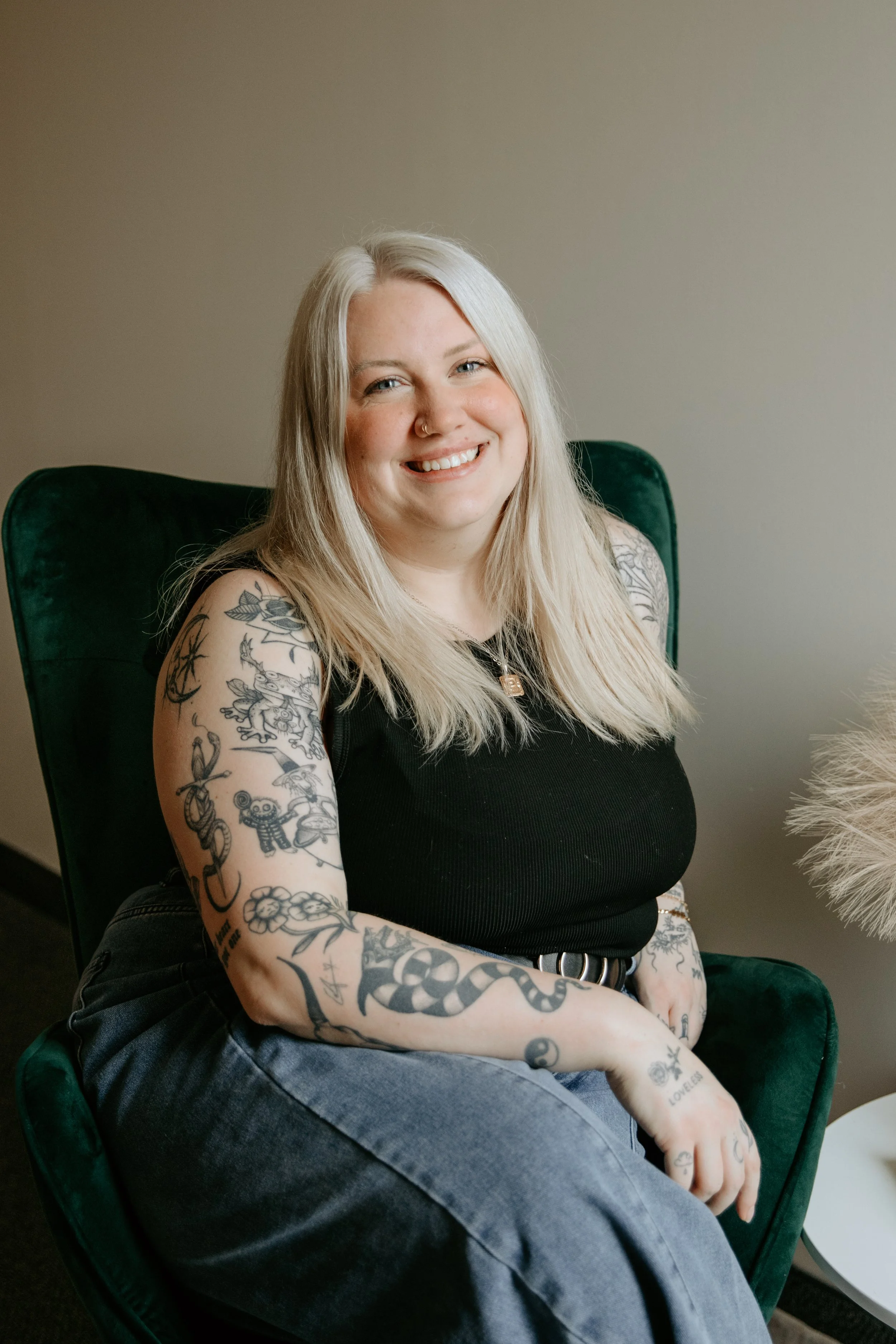 Person with blonde hair sitting on a leather couch, wearing a gray sweater, with tattoos on their arms and hands. A lit candle and lamp are in the background.
