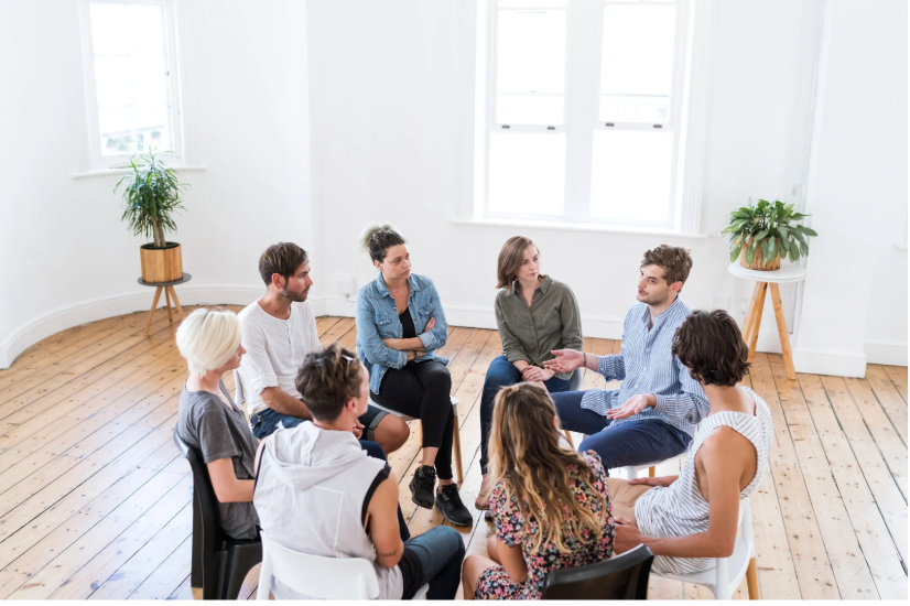 A group of people sitting in a circle during a meeting or discussion in a room with wooden floors and white walls, decorated with potted plants.