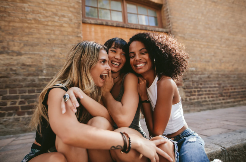 Three women sitting and laughing together in front of a brick building.