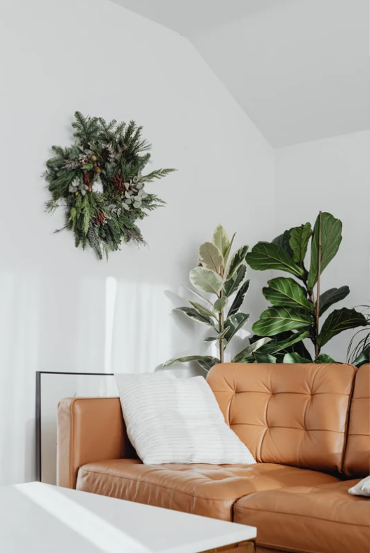 Modern living room with tan leather sofa, white pillow, green plants, and decorative wreath on the wall.