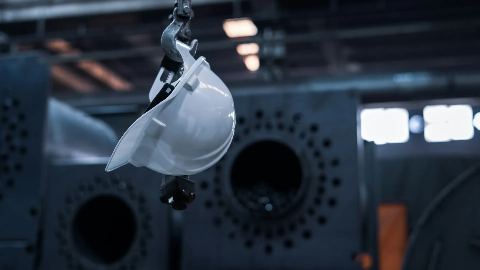 A white safety helmet hanging from a hook in an industrial setting with metal structures and ventilation systems in the background.