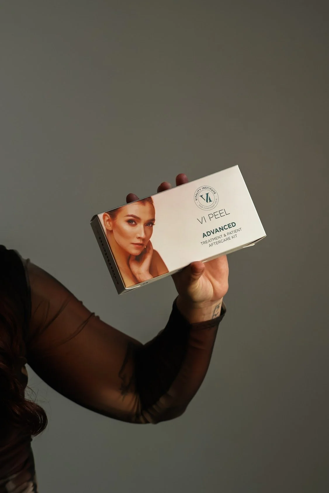 A person wearing a black, sheer, long-sleeve top holds a box of VI Peel Advanced treatment and patient aftercare kit with a photo of a woman on the packaging, against a plain gray background.